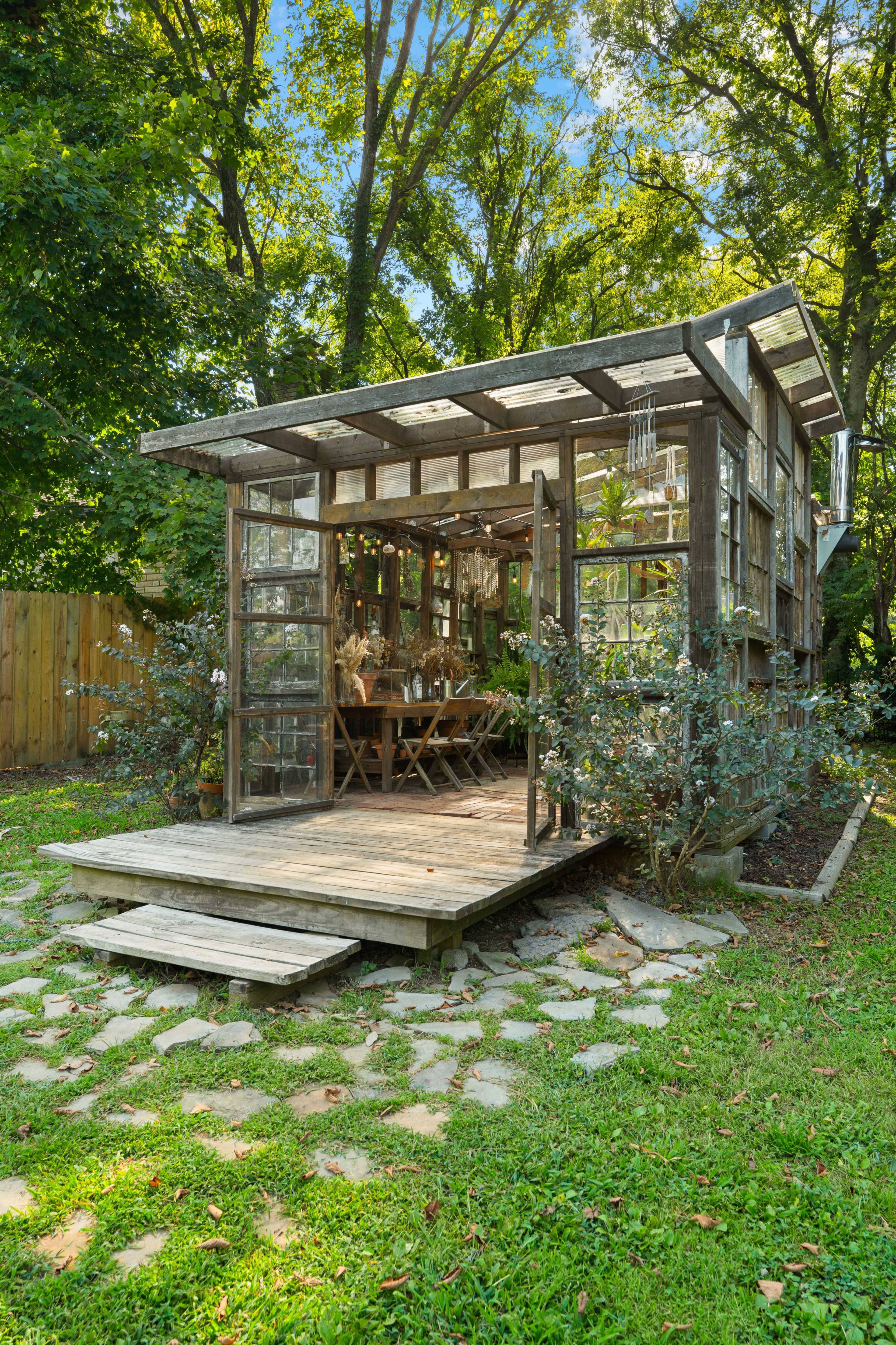 A wooden garden shed with glass walls and an open front is surrounded by lush greenery and stone pathways.
