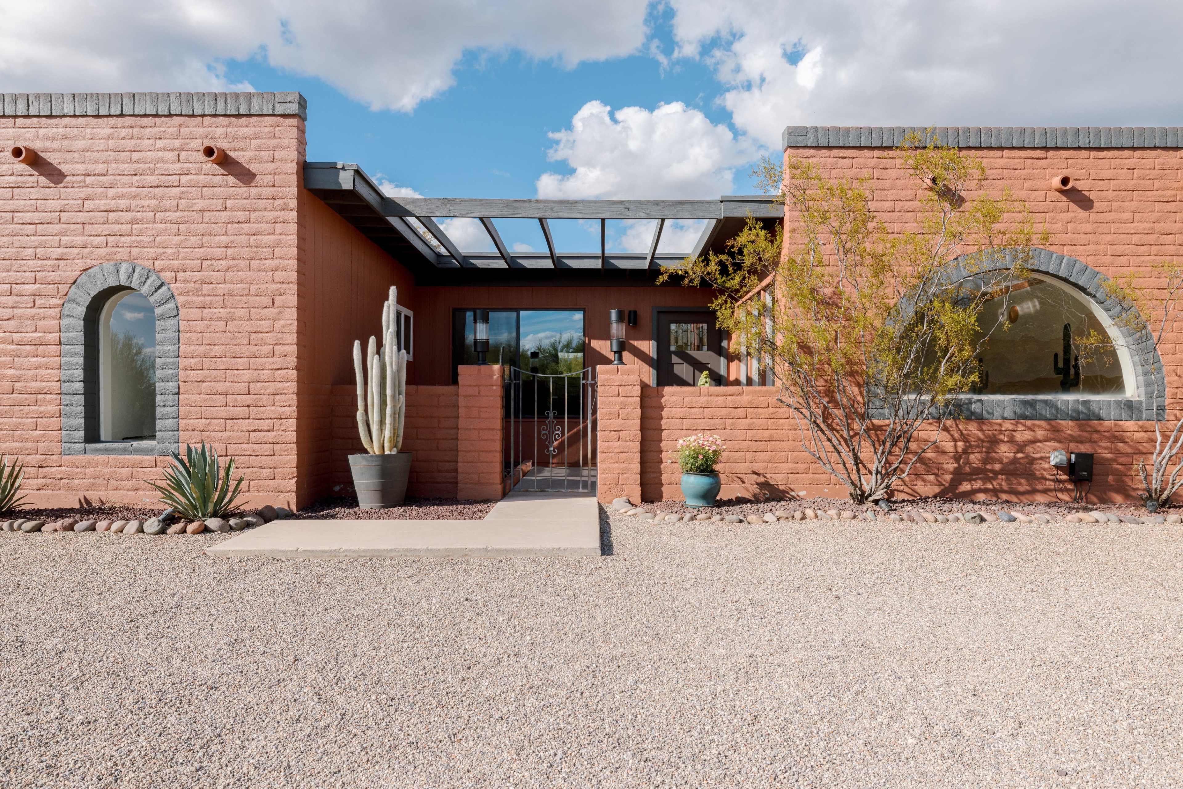 A modern southwestern-style house with a gravel driveway, featuring a red adobe exterior, large windows, and desert landscaping.