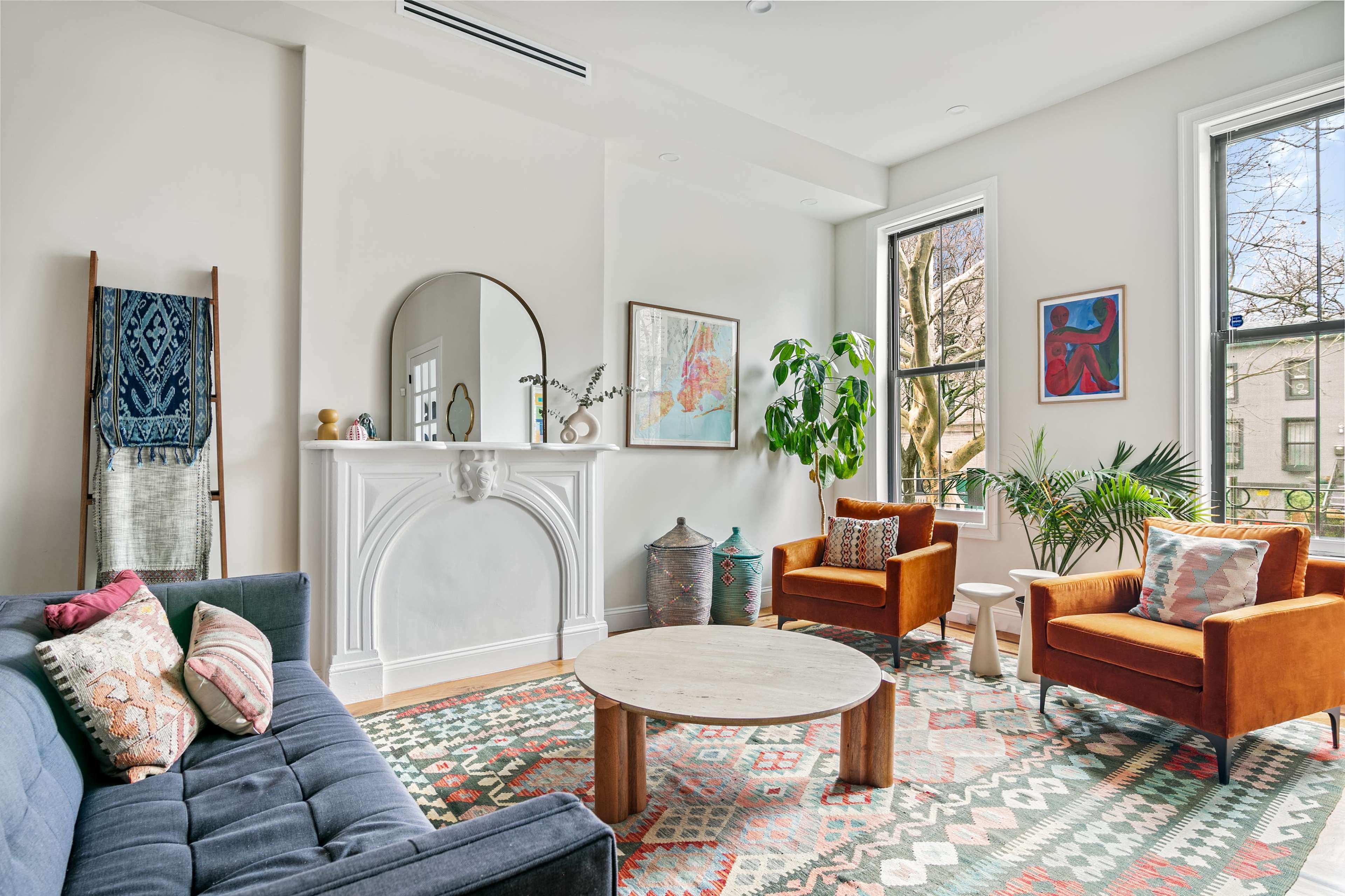 The image shows a stylish living room with a blue sofa, two brown chairs, a round wooden coffee table, and large windows letting in natural light.
