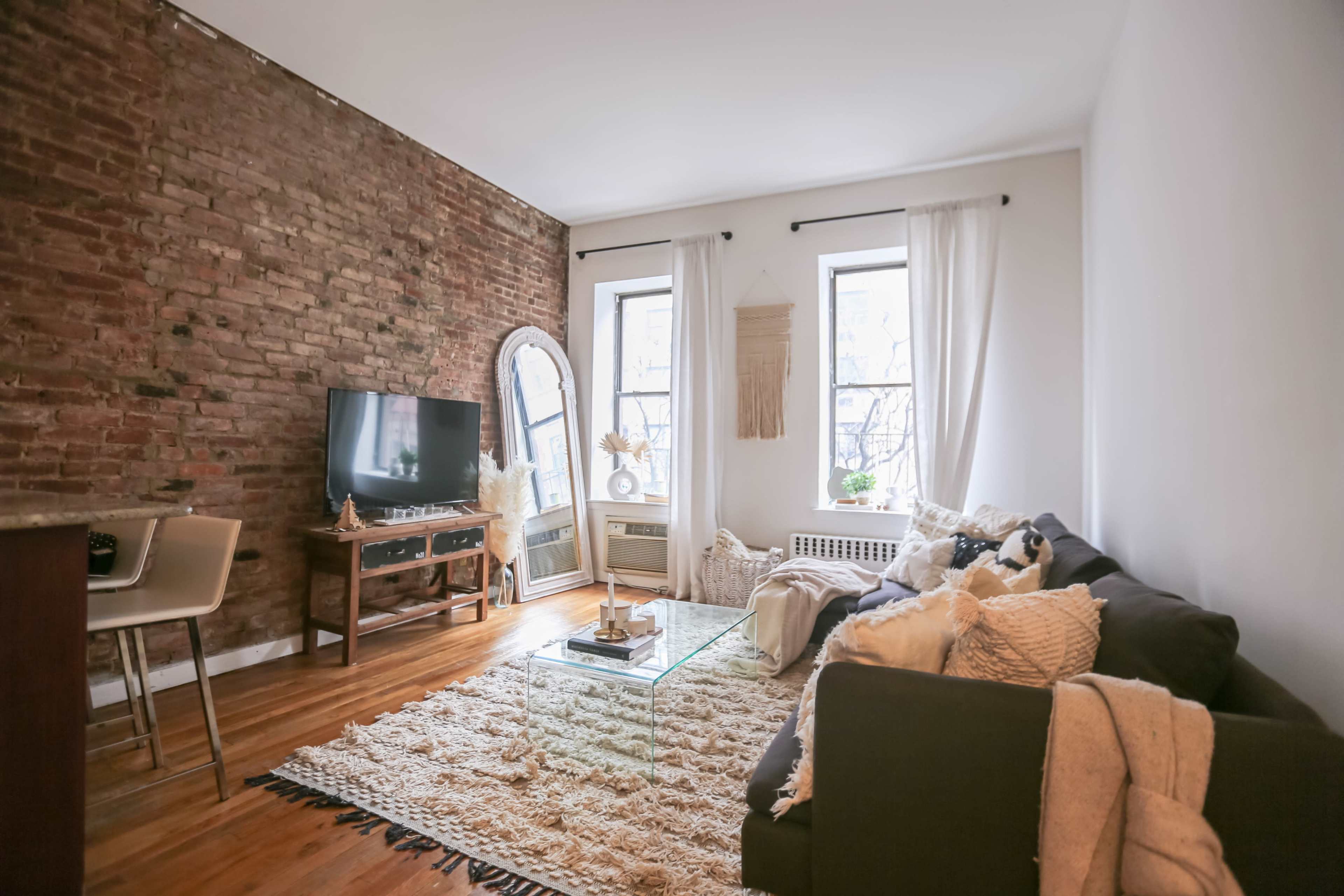 A cozy living room features exposed brick walls, large windows, a TV stand, and a plush rug with a sofa adorned with cushions.