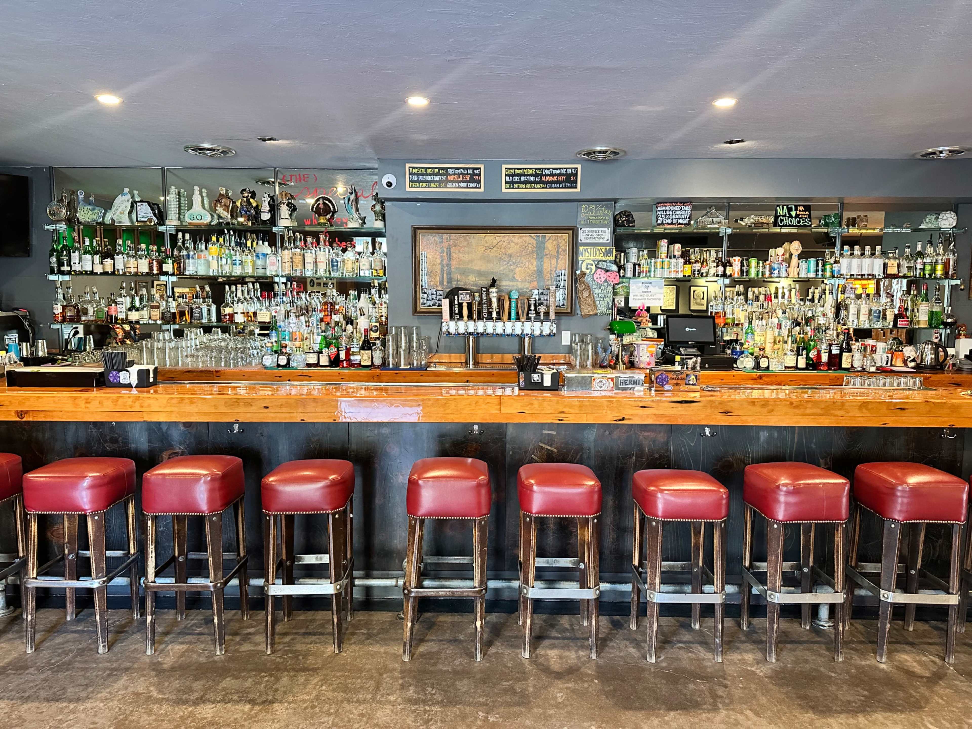 The image shows a neatly arranged bar with a wooden countertop, red leather stools, and shelves filled with various bottles of alcohol and drinkware.