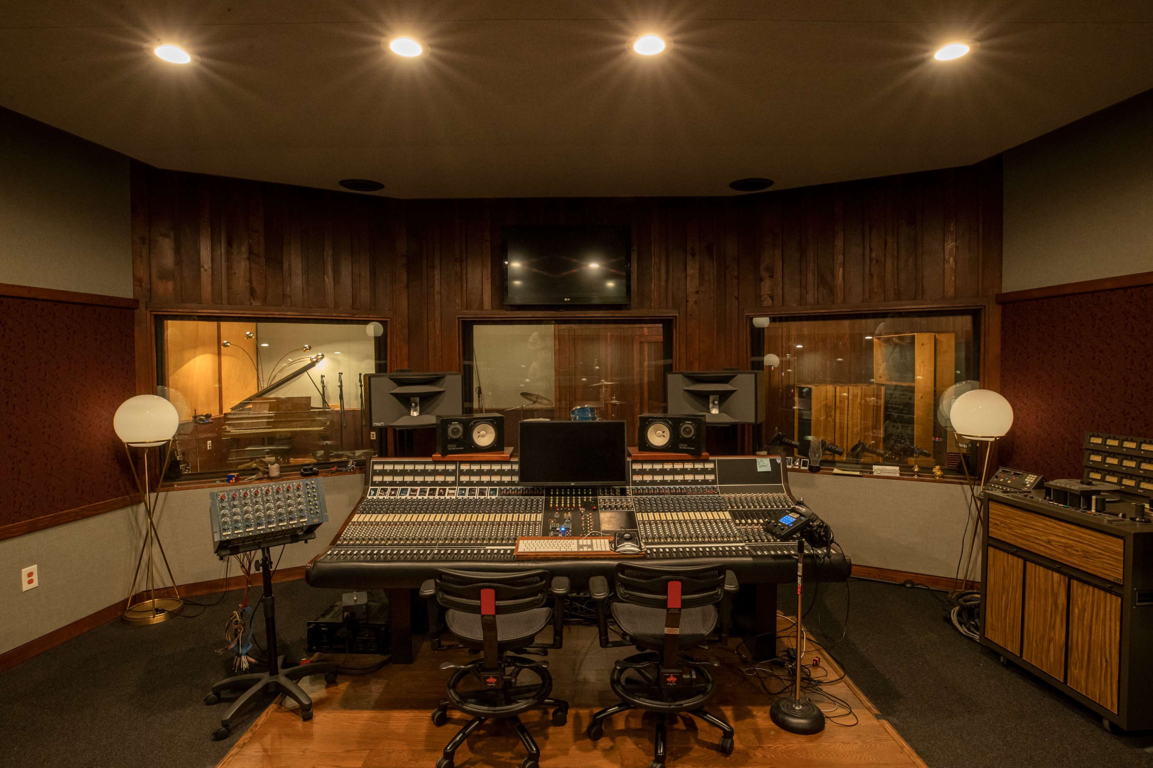 A recording studio with a large mixing console, monitors, and soundproof glass windows overlooking a sound booth.