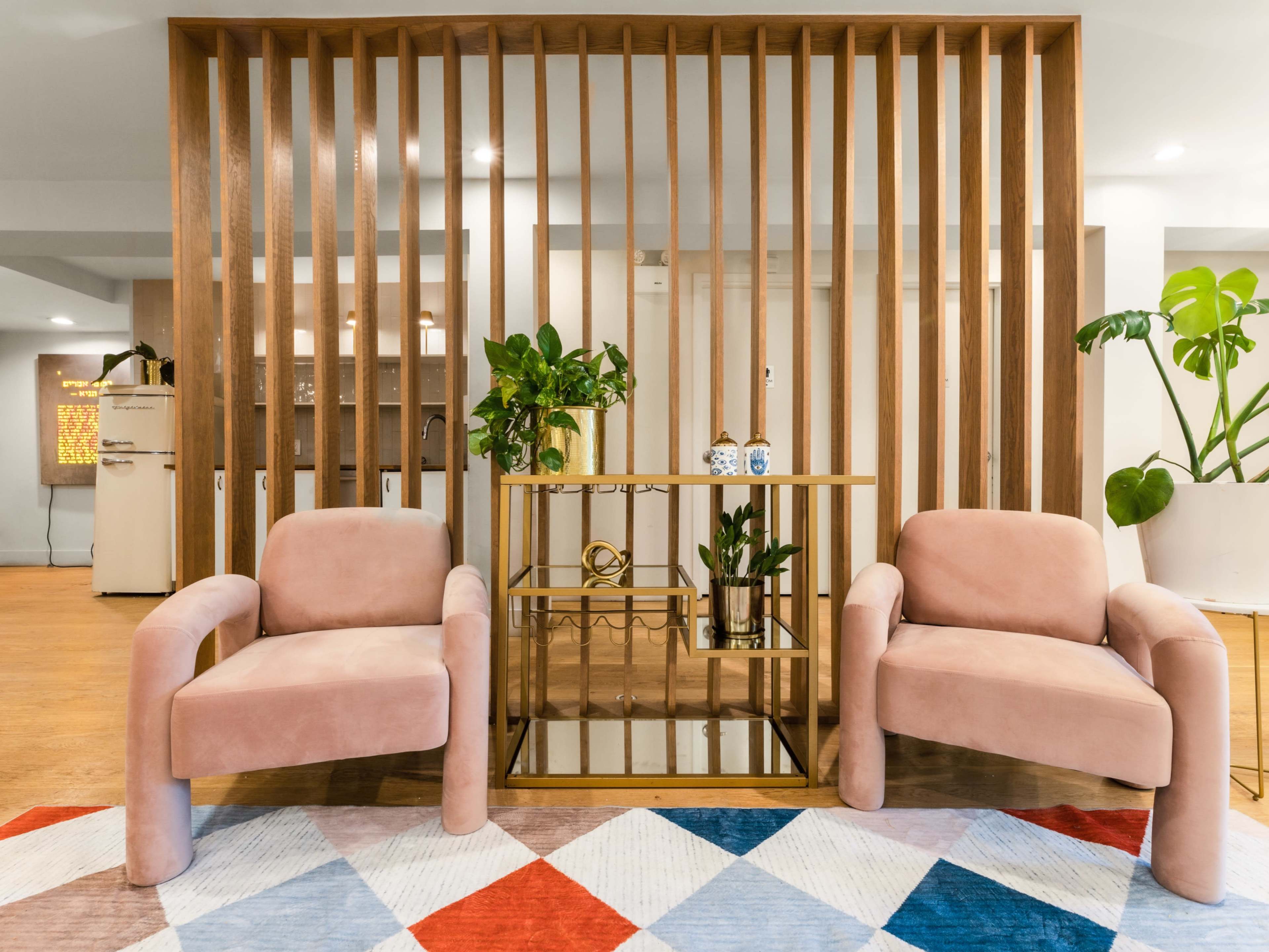 The image shows a modern living space featuring two pink upholstered chairs facing a wooden divider with decorative plants and a metallic coffee table.