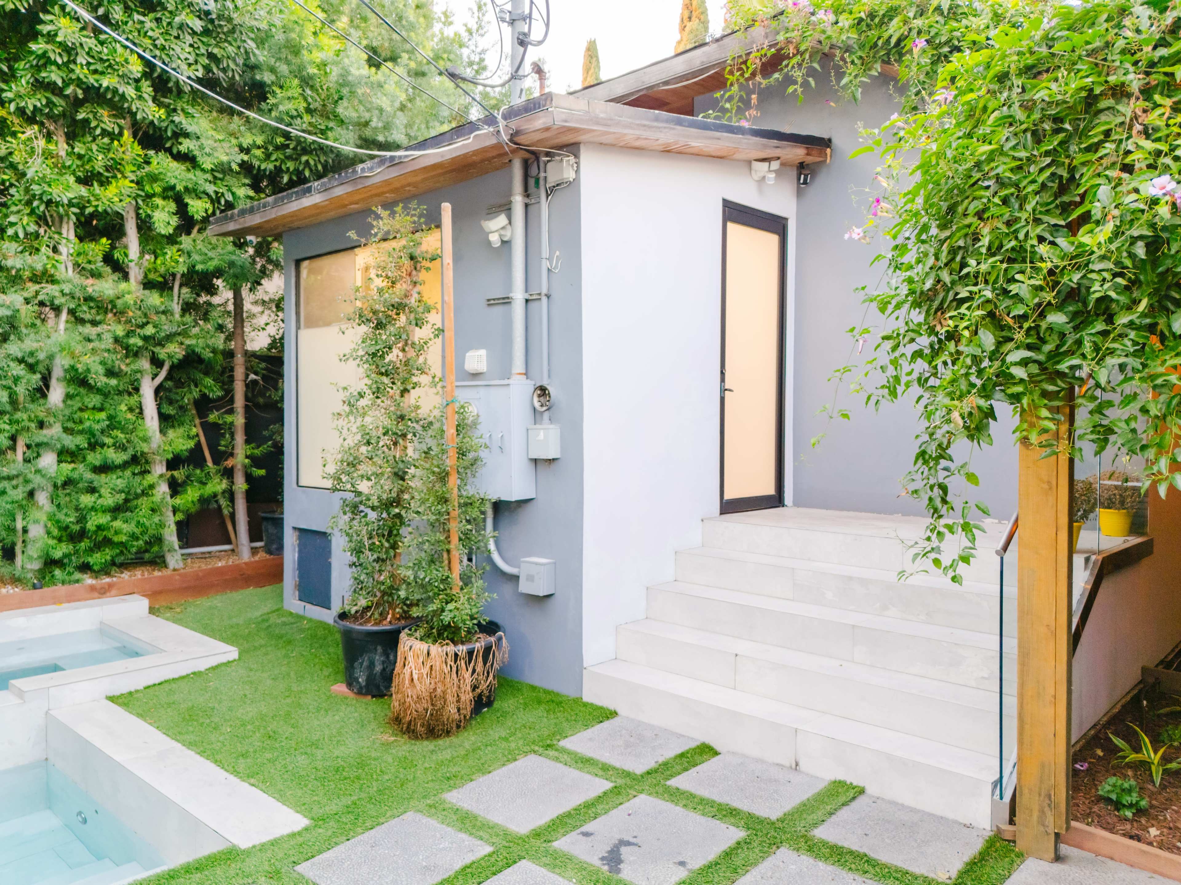 The image shows a modern outdoor area featuring a small building with a staircase leading up to a door, surrounded by greenery and a small pool adjacent to the structure.