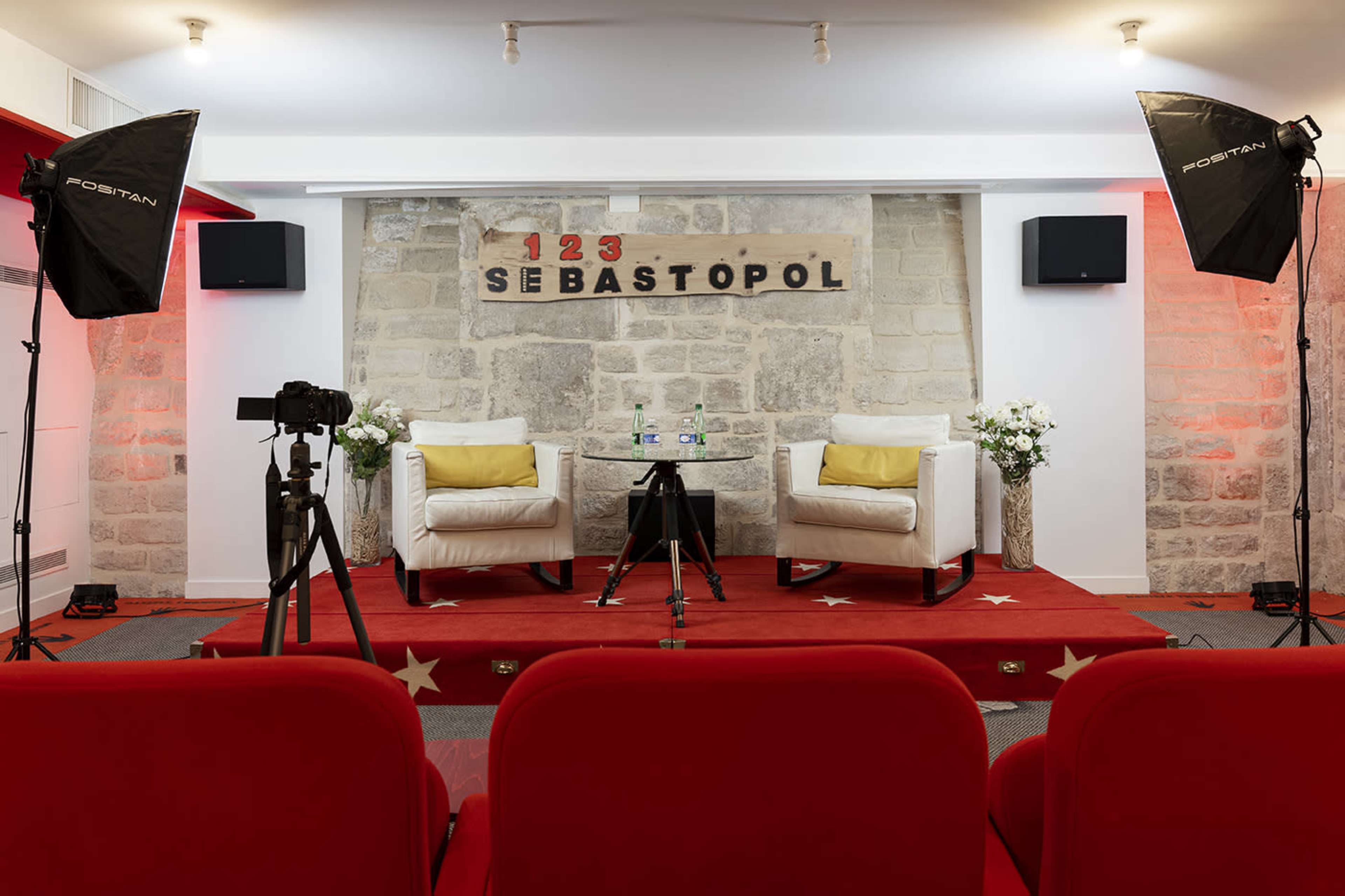 A staged interview set with two white armchairs, a small black table, cameras, and lighting equipment, arranged in front of a textured stone wall.