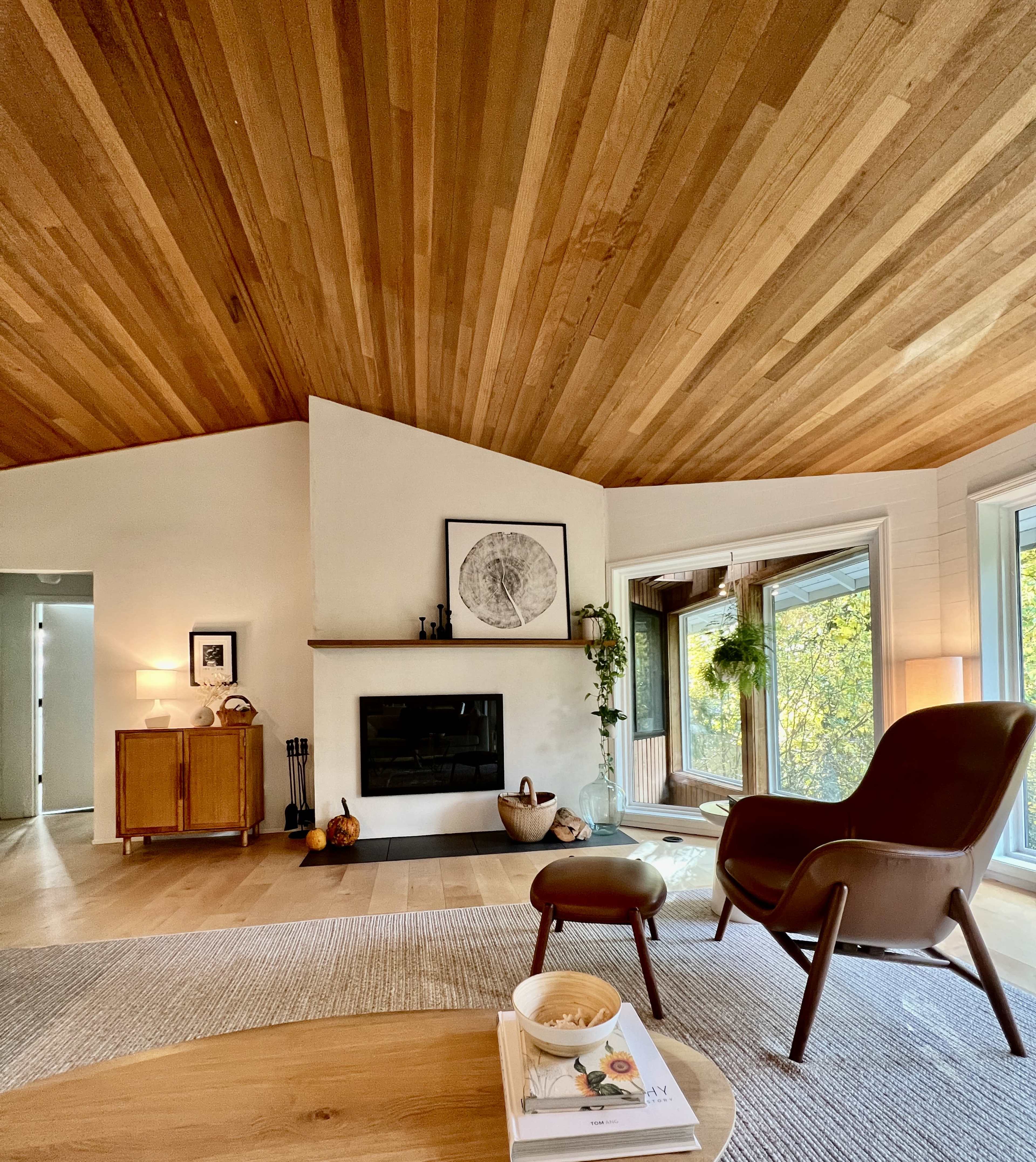 A modern living room with wooden ceiling beams, a wall-mounted fireplace, and large windows featuring a view of greenery outside.
