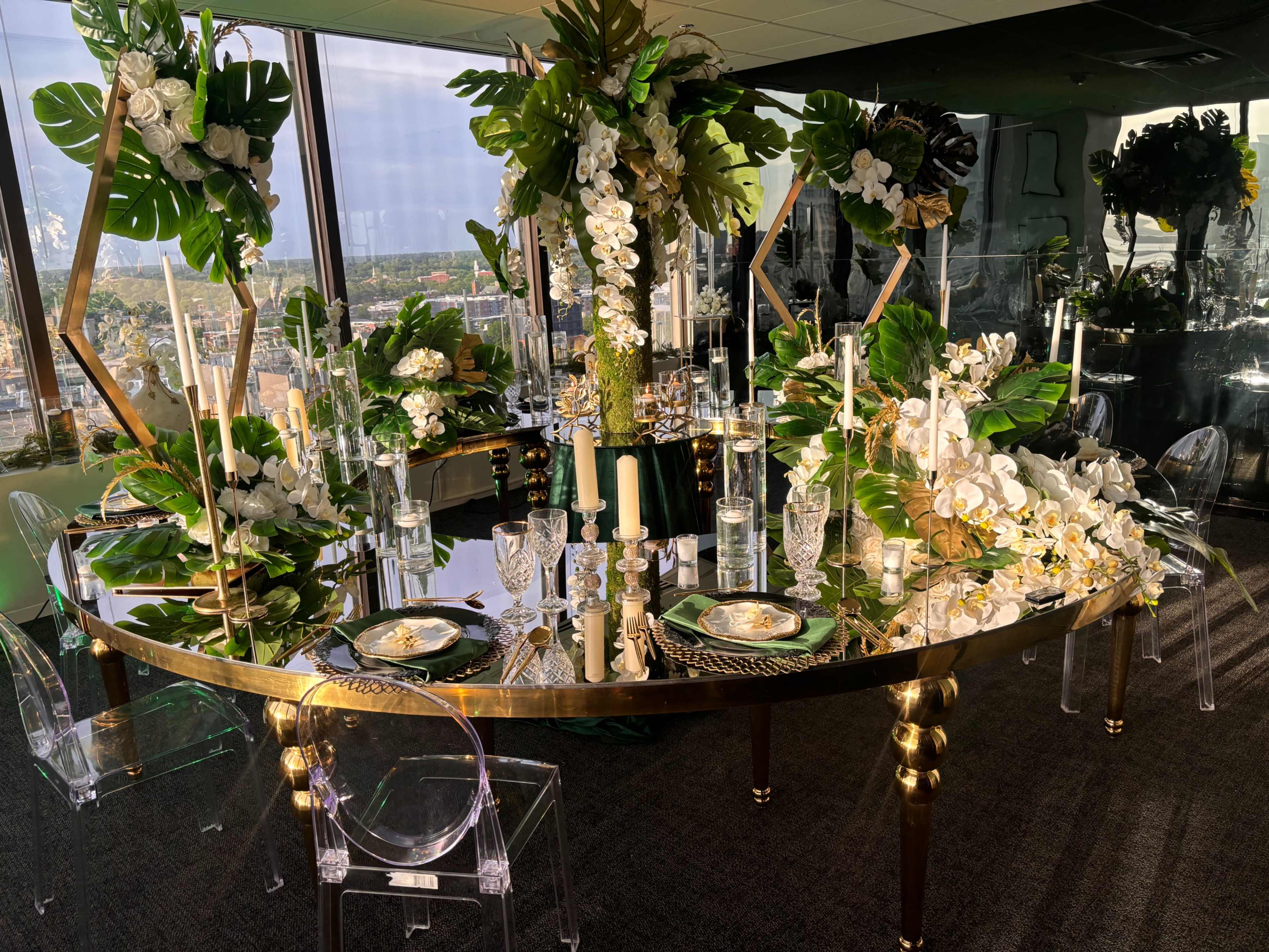 A round dining table is elegantly set with floral arrangements, candles, and glassware against a panoramic city view.