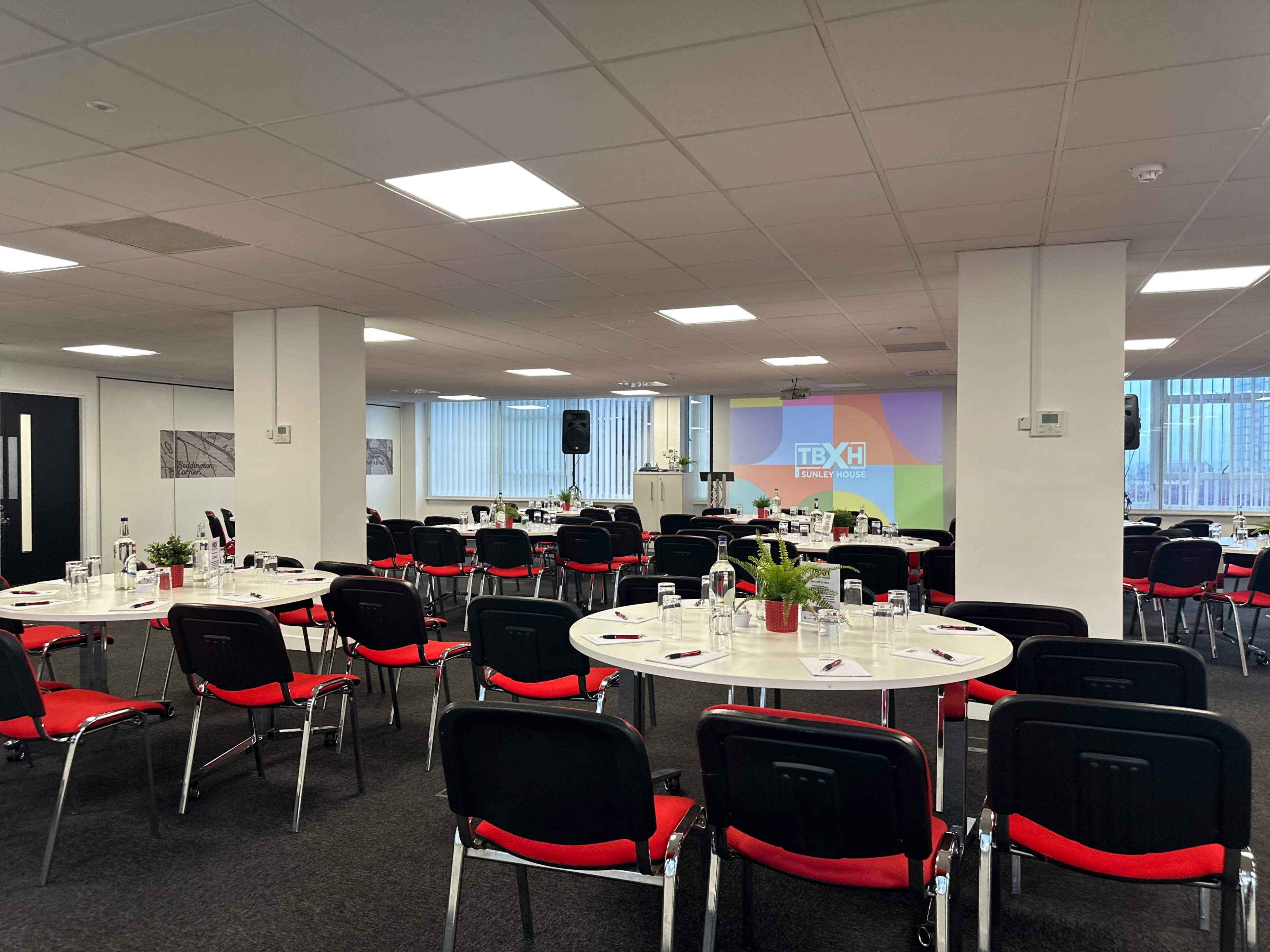 A conference room is set up with circular tables, red chairs, and a projector screen displaying a logo.
