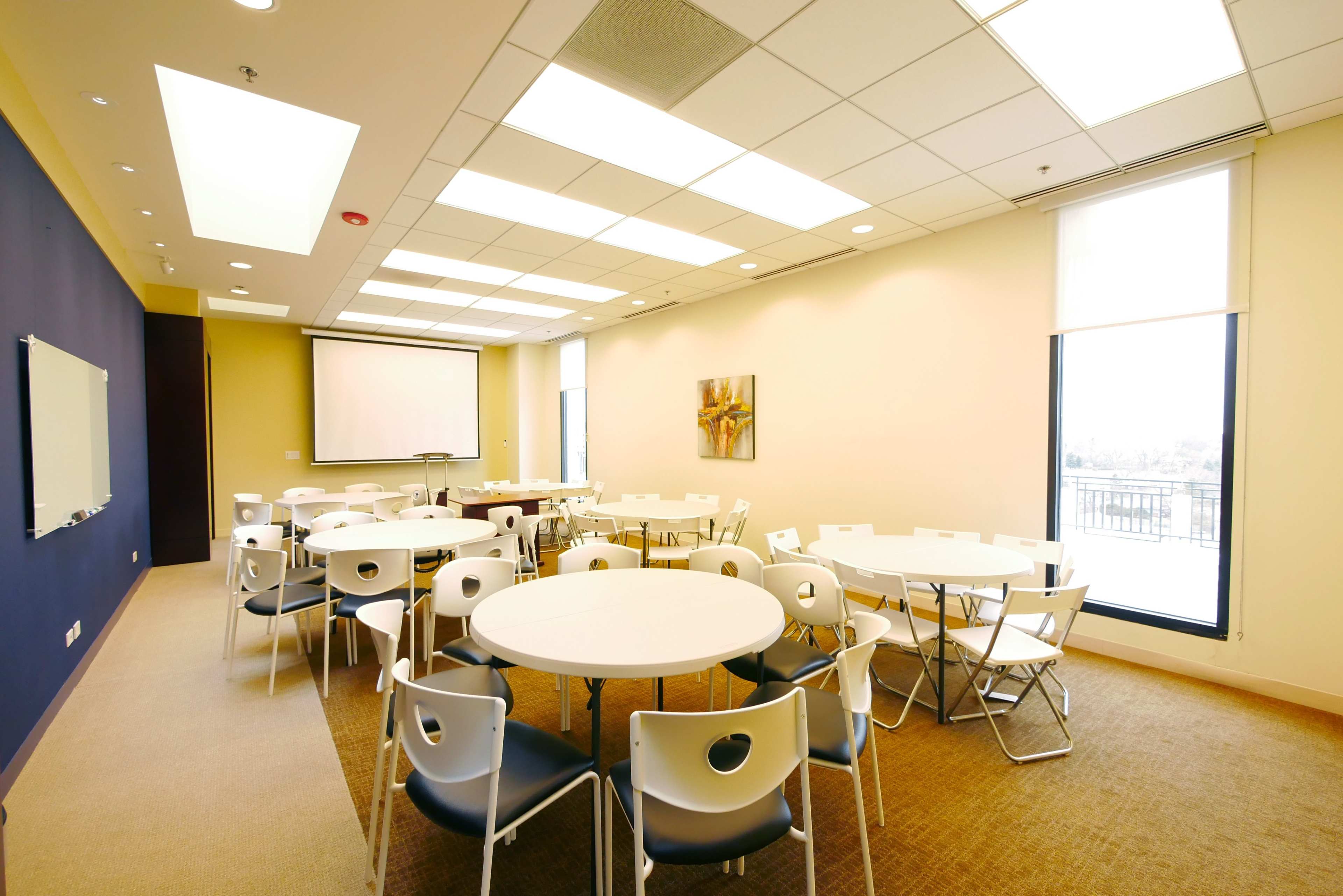 The image shows a modern meeting room with several round tables and chairs, equipped with a projector screen and windows offering natural light.