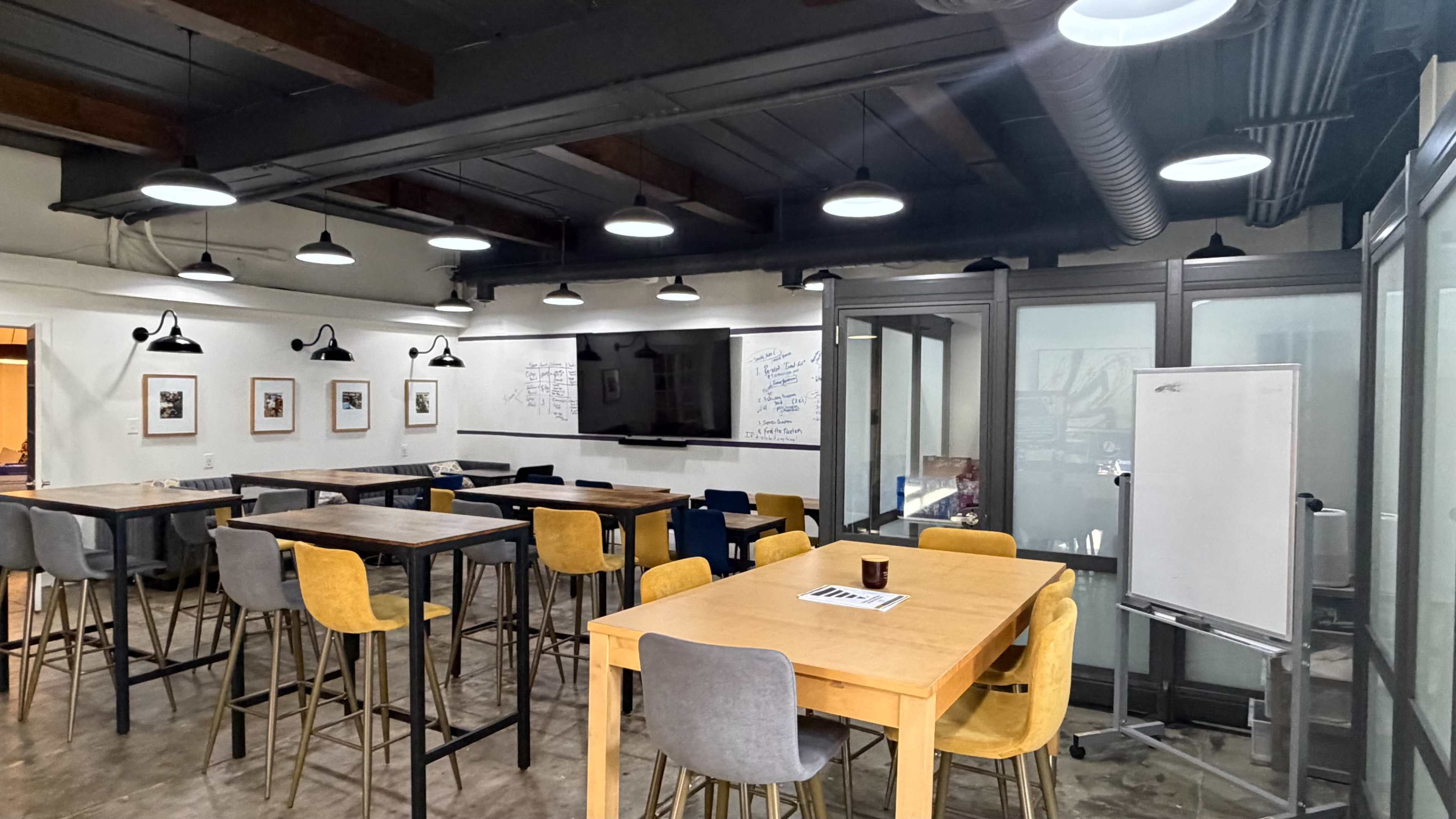 The image shows a modern office space with several tall tables surrounded by chairs, a large screen on one wall, and whiteboards with writing.