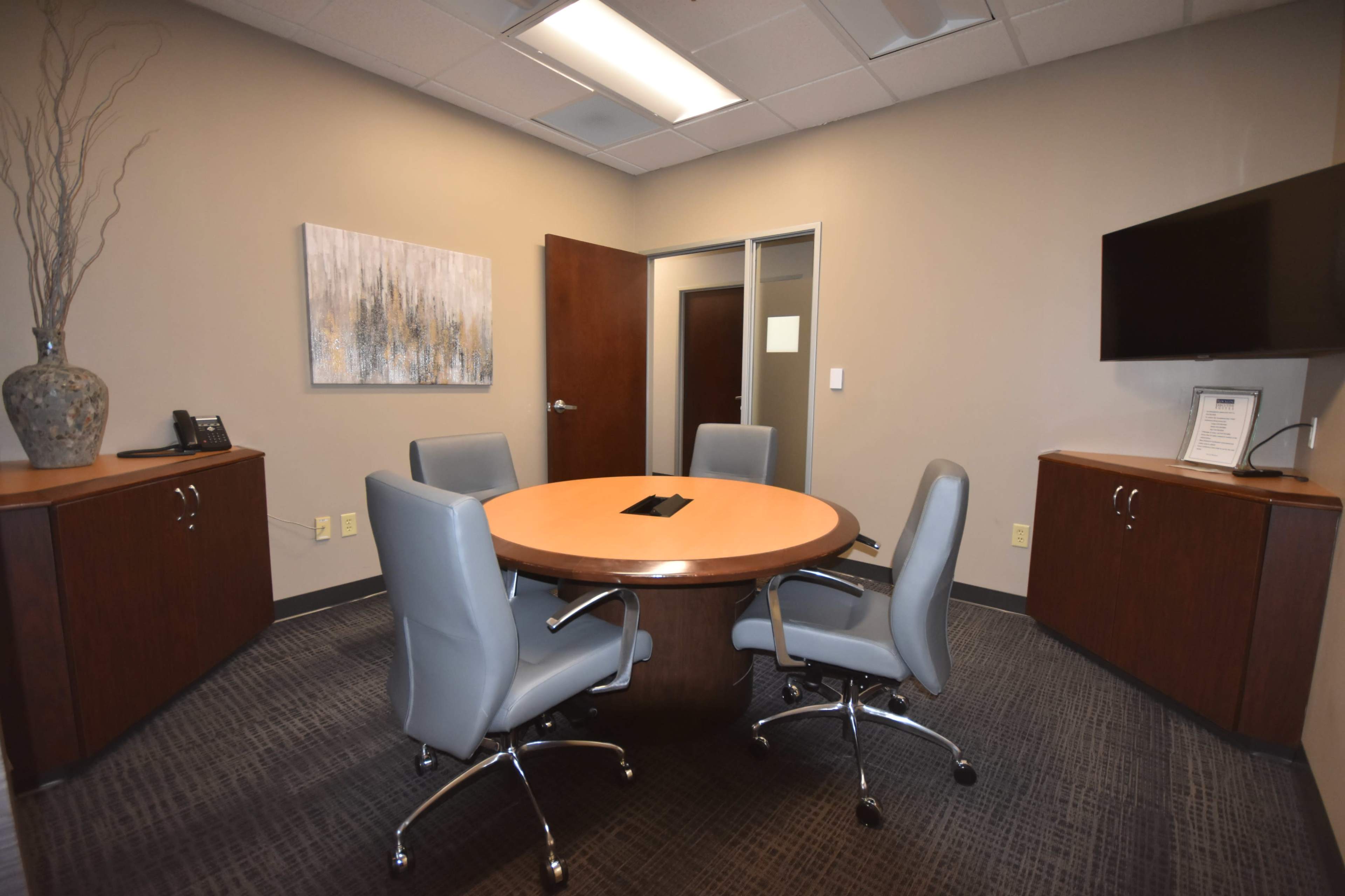 A corporate meeting room featuring a circular wooden table surrounded by four gray chairs, with a television mounted on the wall and cabinets on either side.