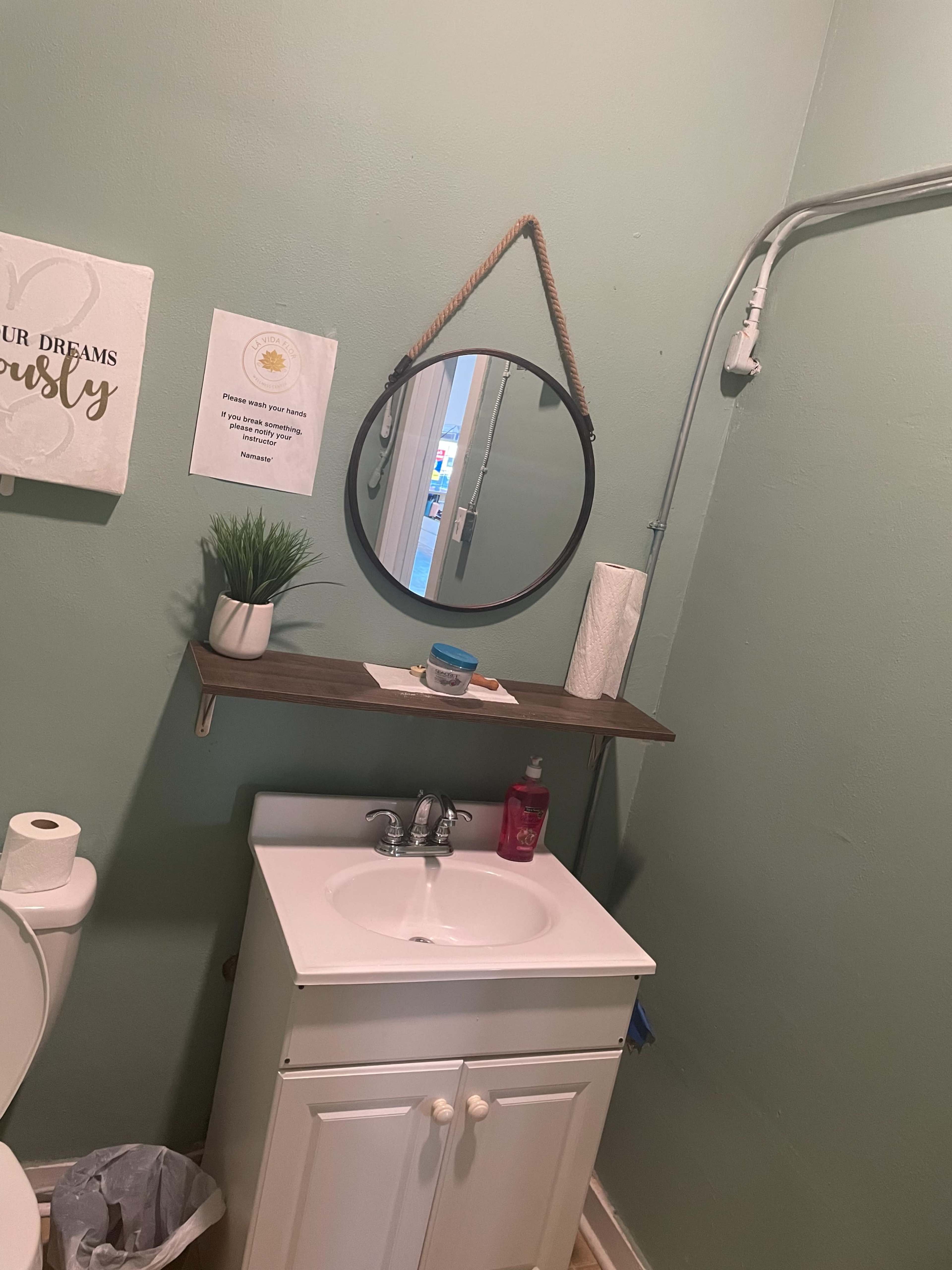 A small bathroom with a circular mirror above a sink, a shelf with a plant, and a notice on the wall.