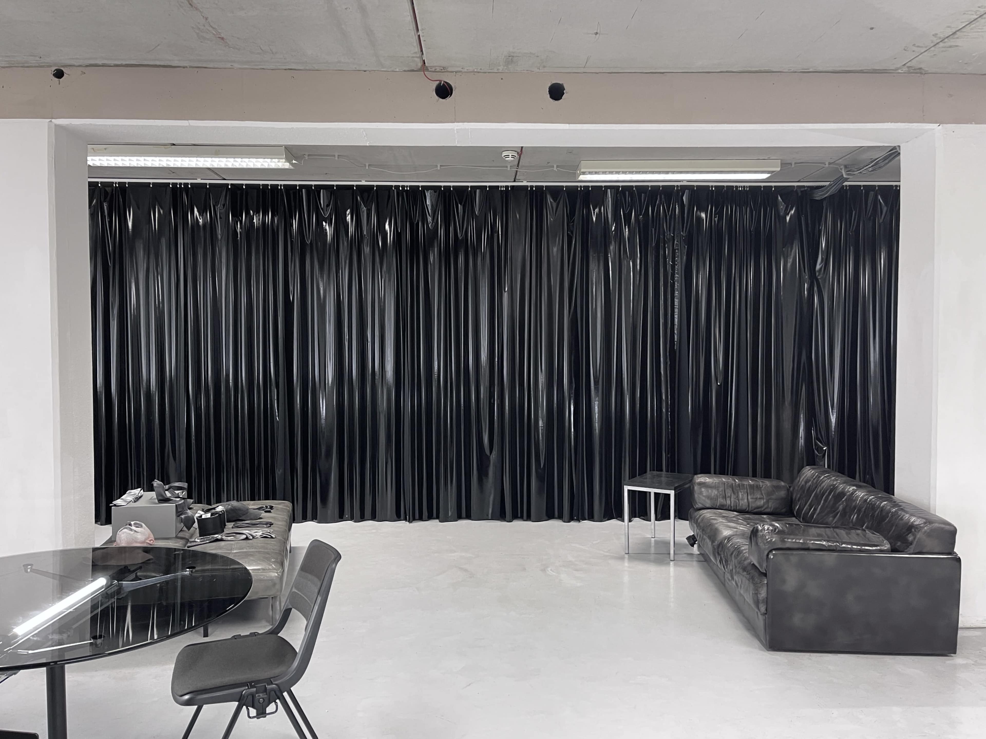 A black curtain lines the wall of a minimalistic room featuring a glass dining table, several chairs, and a leather sofa.
