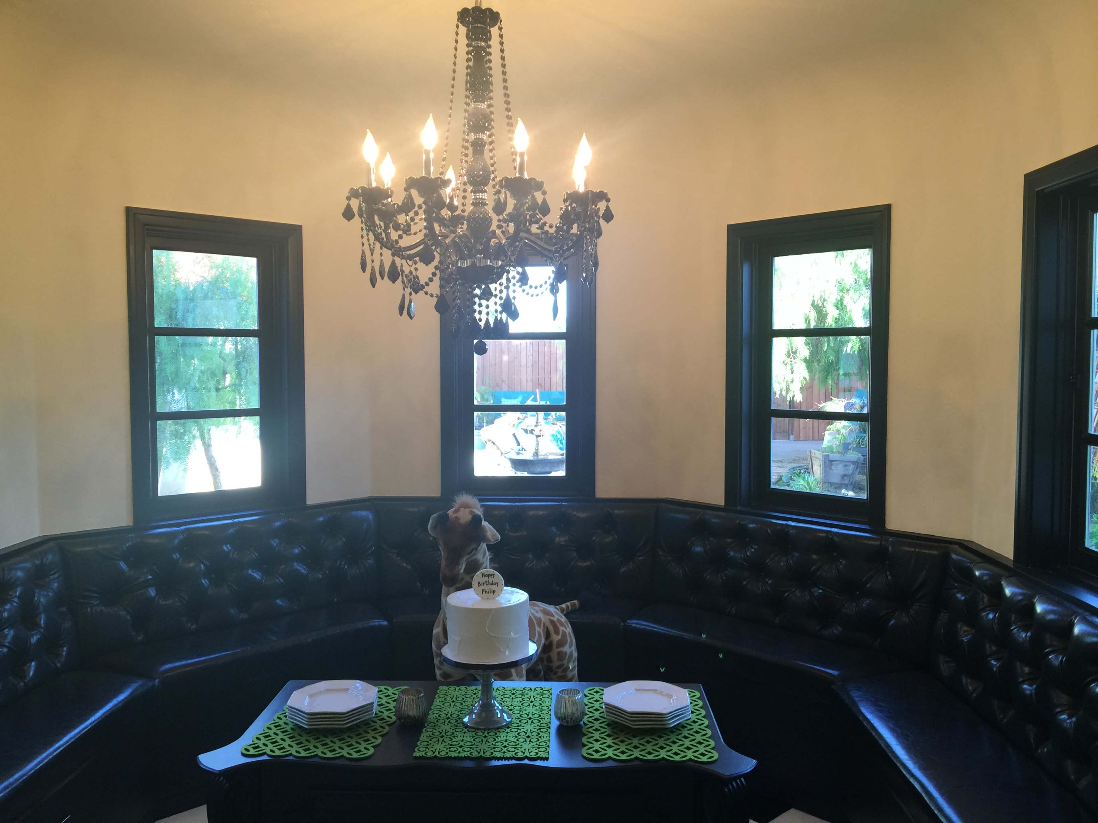 A dining area features a black leather banquette, a decorative chandelier, and a cake stand on a table with green accents.