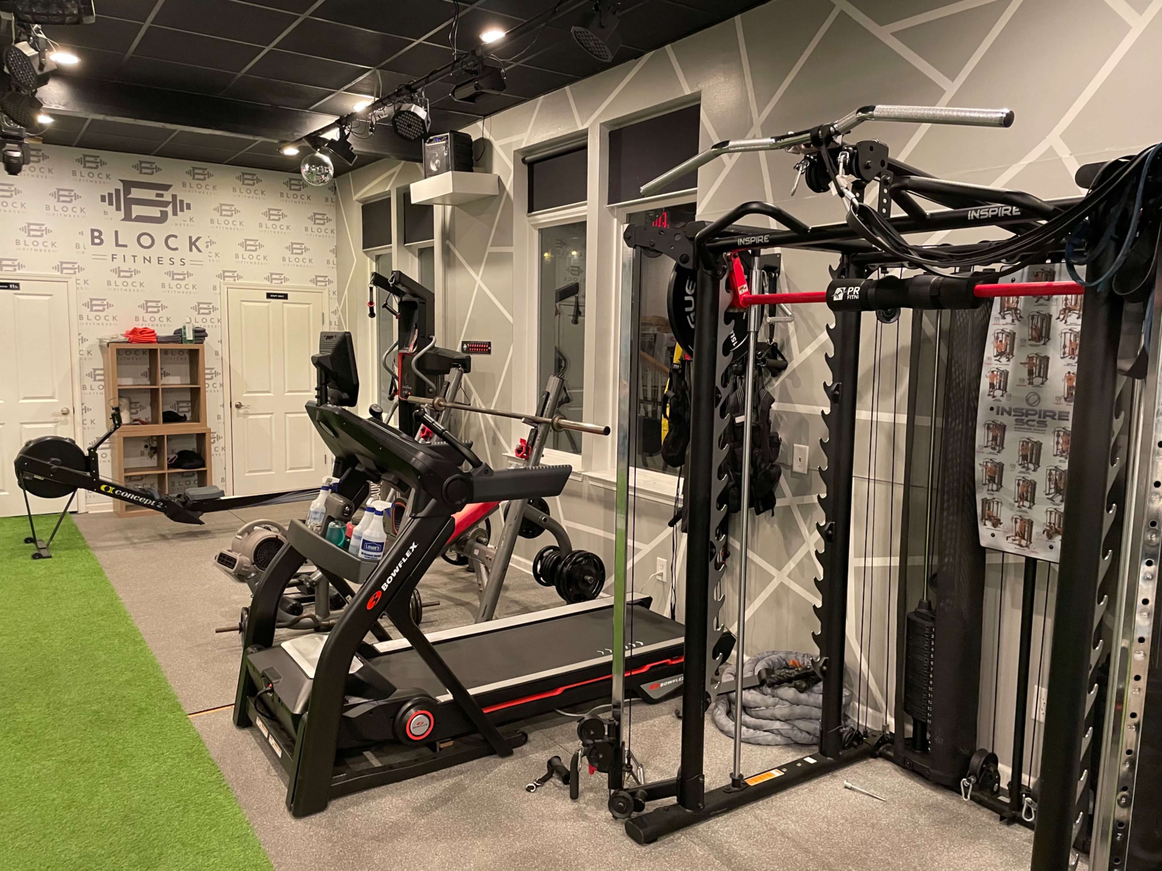 A fitness room equipped with a treadmill, strength training machine, exercise bike, and rowing machine, along with a green turf area.