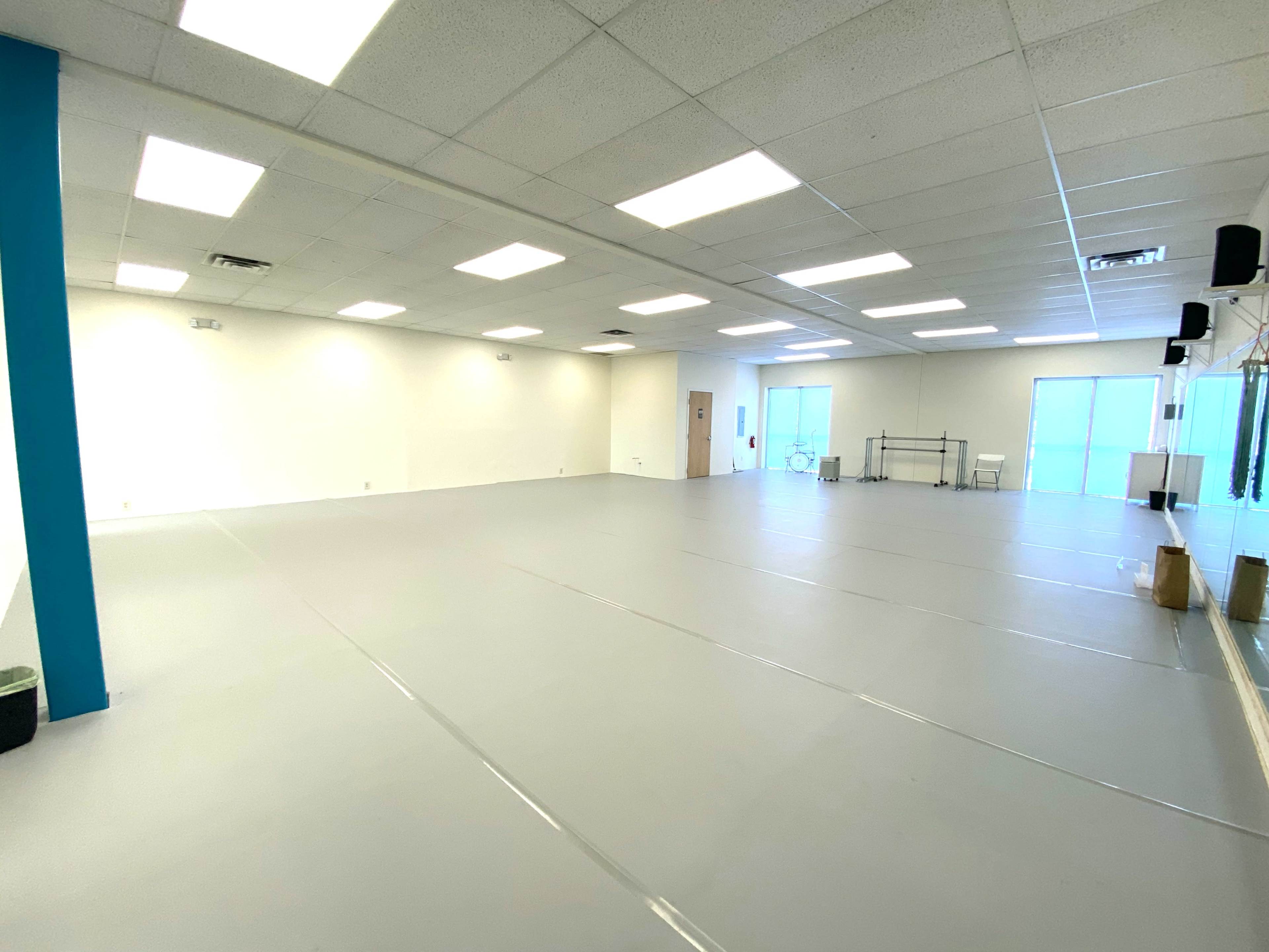 A large, empty studio space with grey flooring and white walls, featuring mirrors and bright overhead lighting.