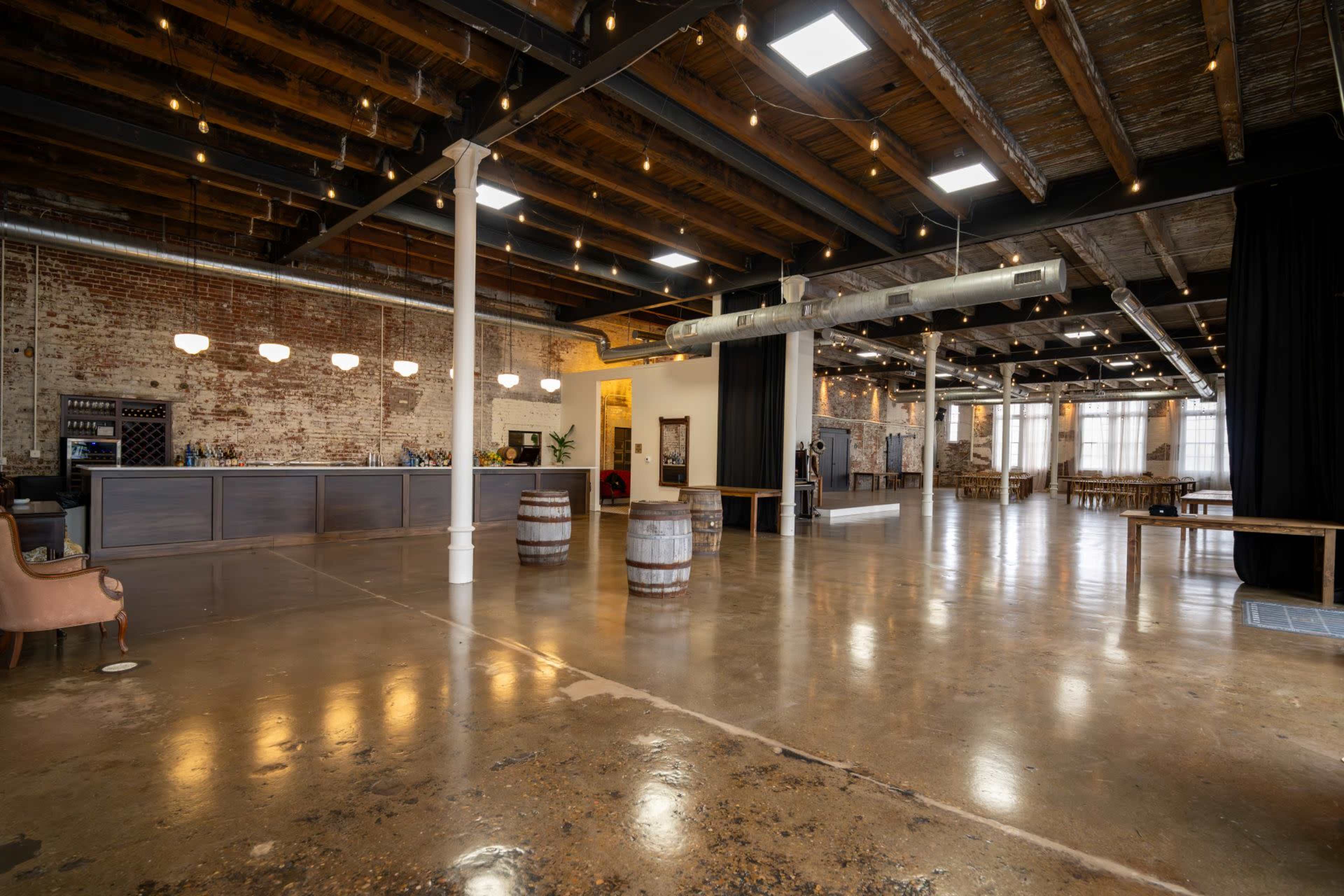 The image shows an empty rustic event space with exposed brick walls, wooden beams, and polished concrete floors, featuring a reception area with a bar and several barrels.