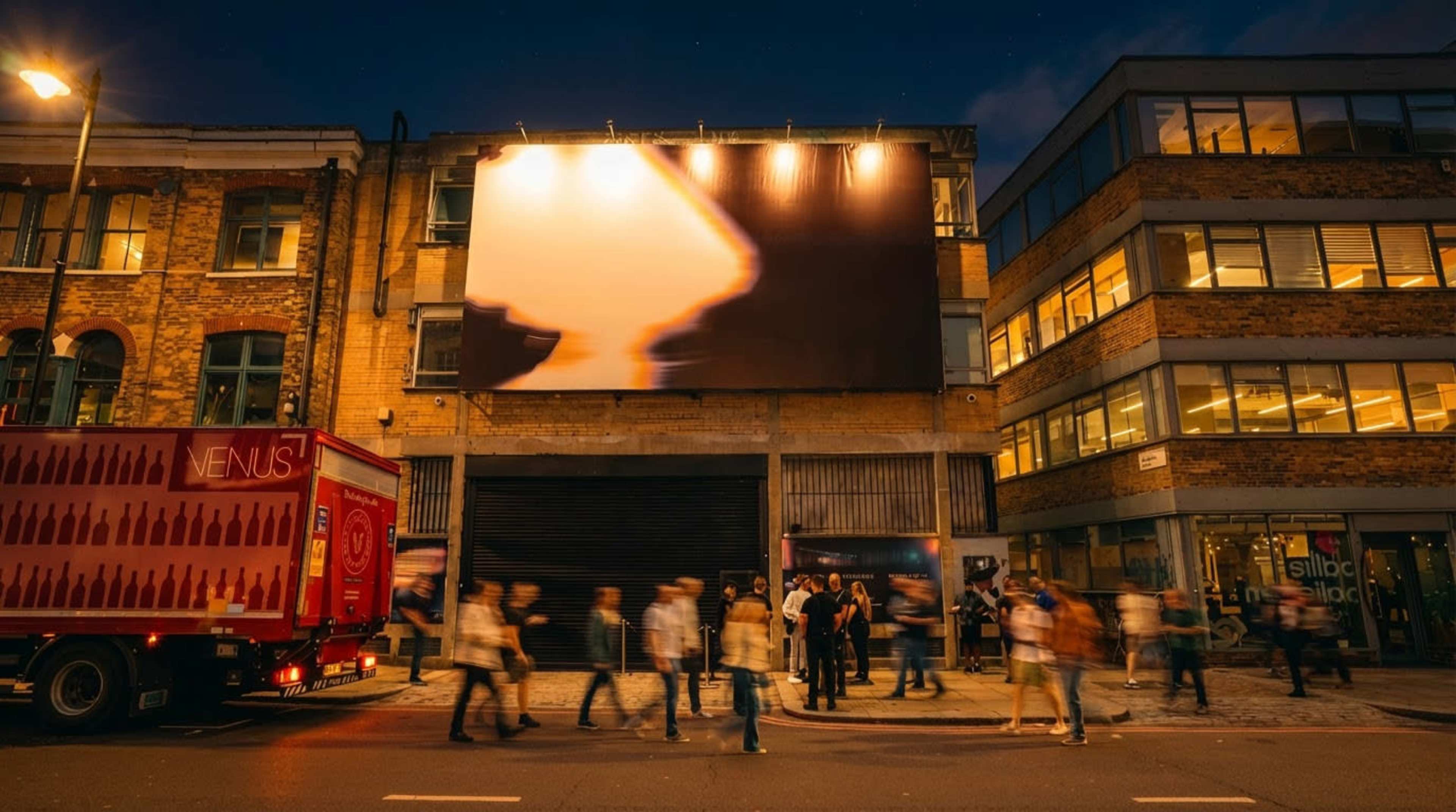A large digital billboard illuminates a building front while pedestrians walk along the street in an urban setting at dusk.