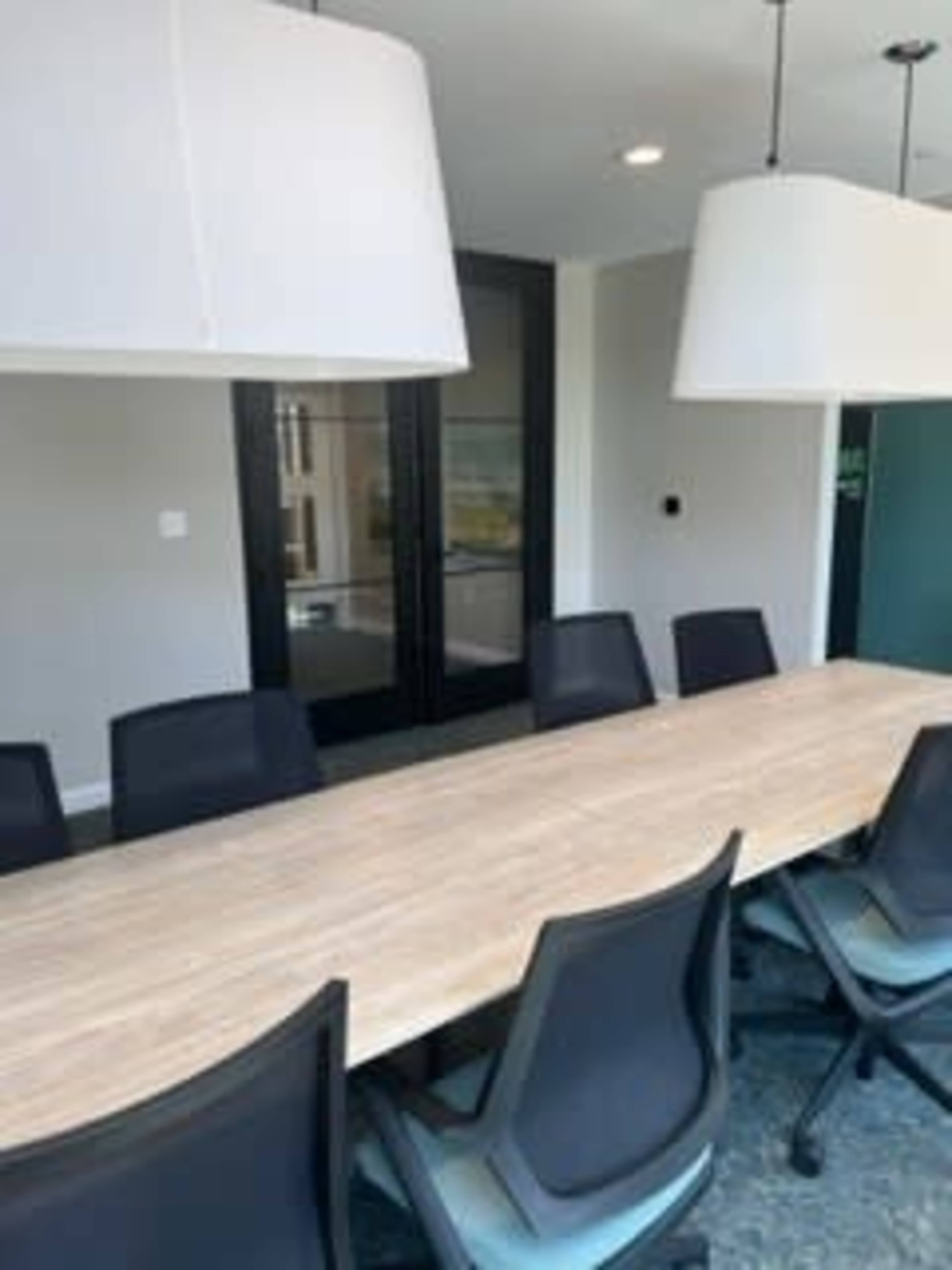 A long wooden conference table with black ergonomic chairs is situated in a modern meeting room, featuring large pendant lights and glass doors.