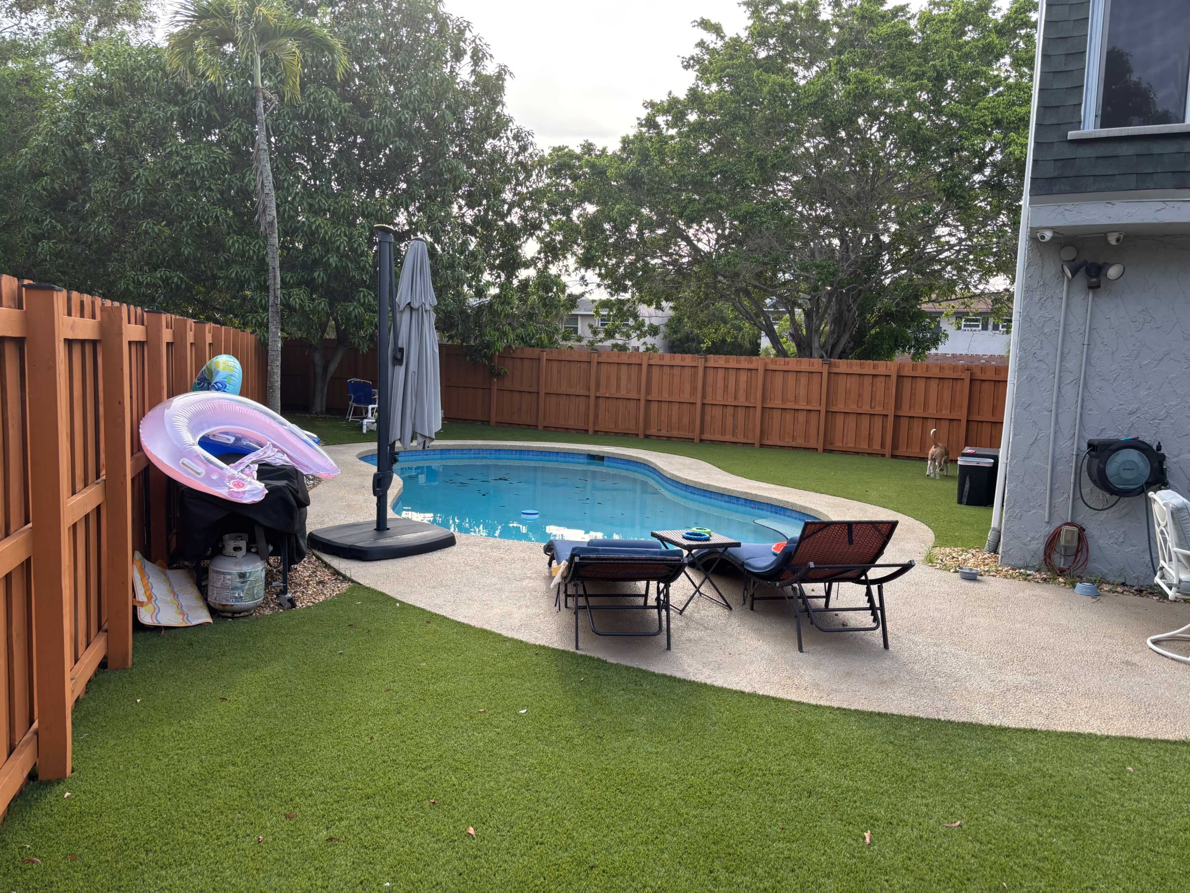 The image shows a backyard pool area surrounded by a low wooden fence, featuring lounge chairs and a floating pool toy.
