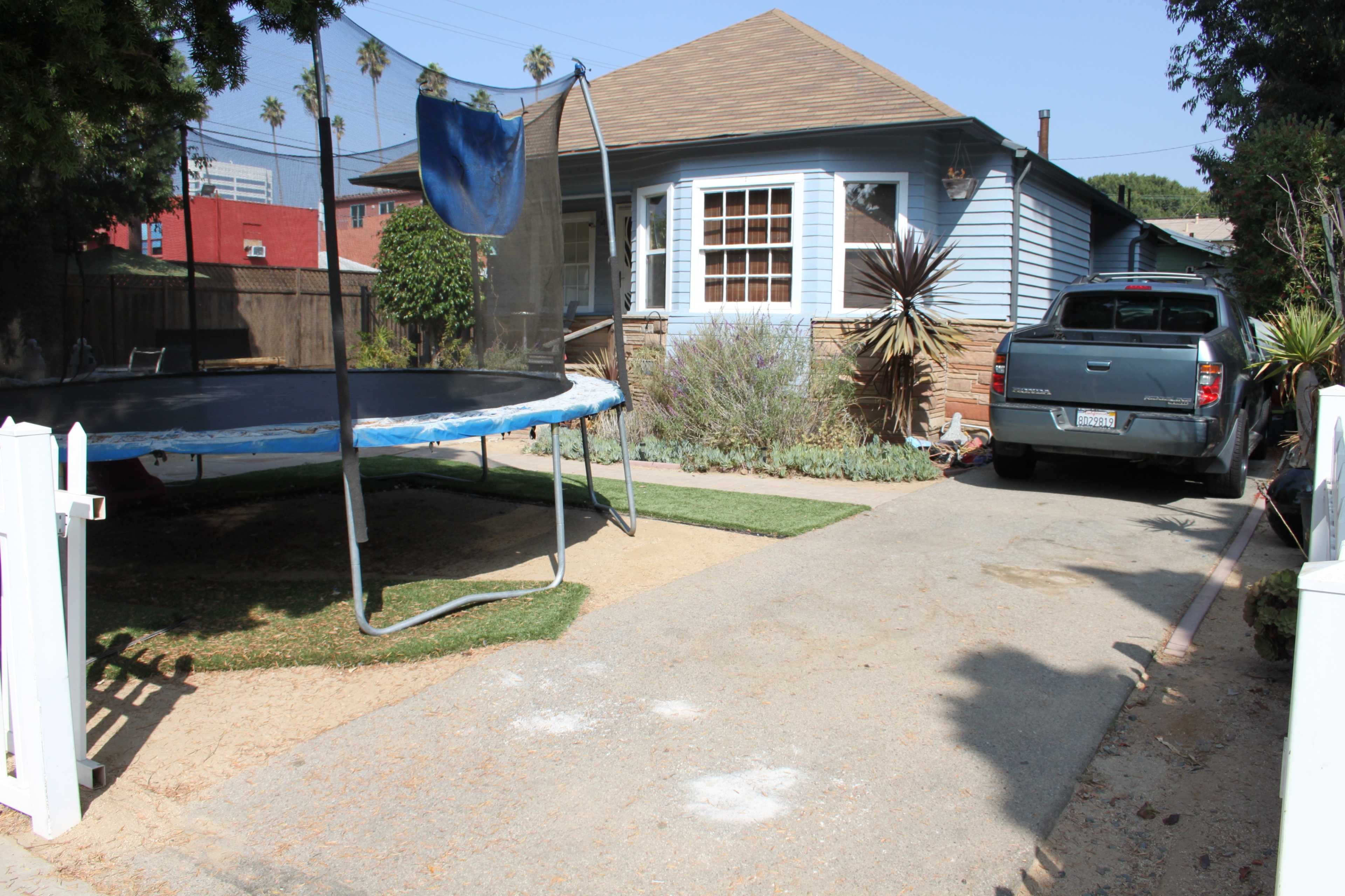 A trampoline stands next to a gravel driveway alongside a house with a blue exterior and a parked vehicle.