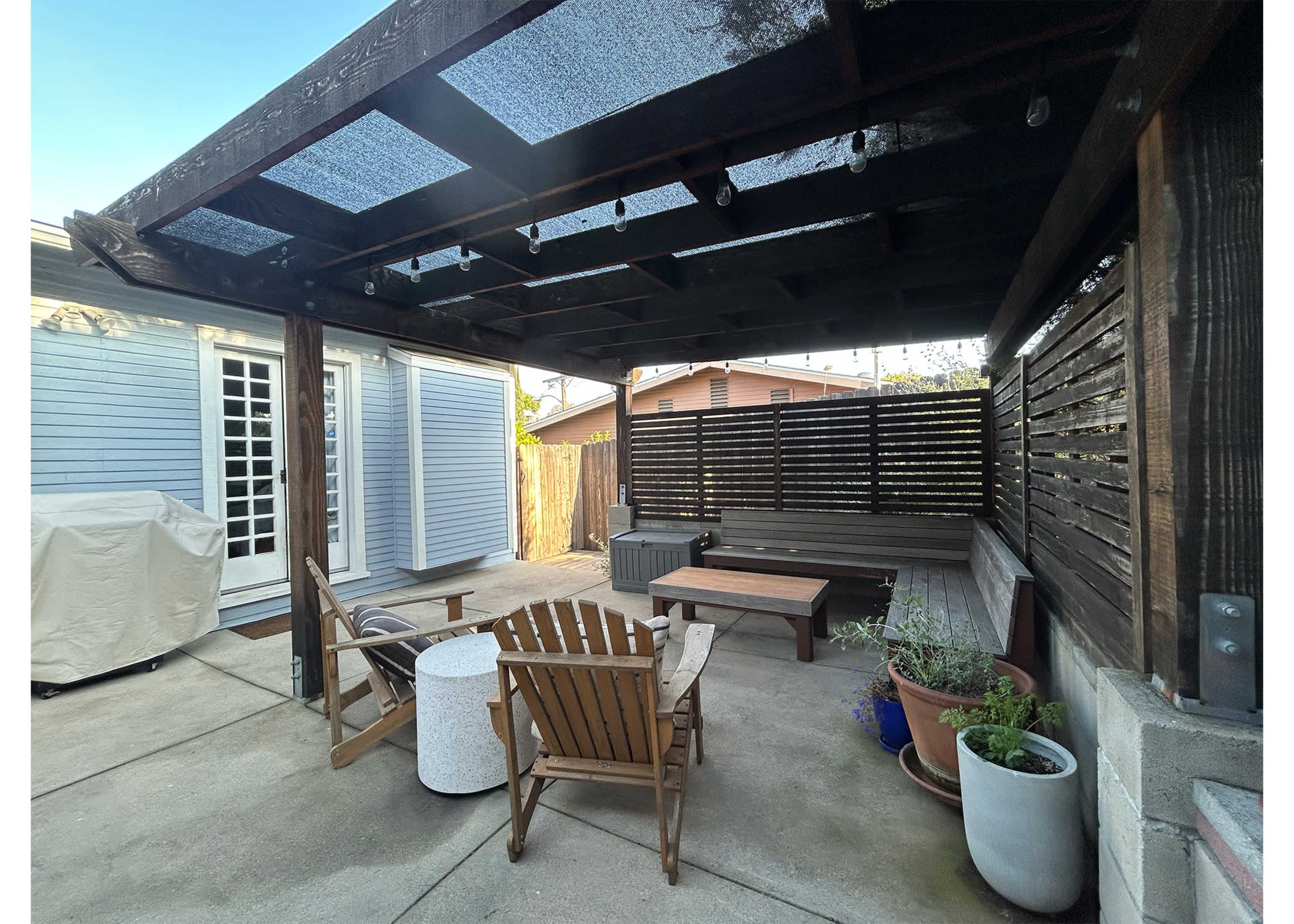The image shows a shaded patio area with wooden chairs, a table, and potted plants, surrounded by a wooden fence and a house in the background.