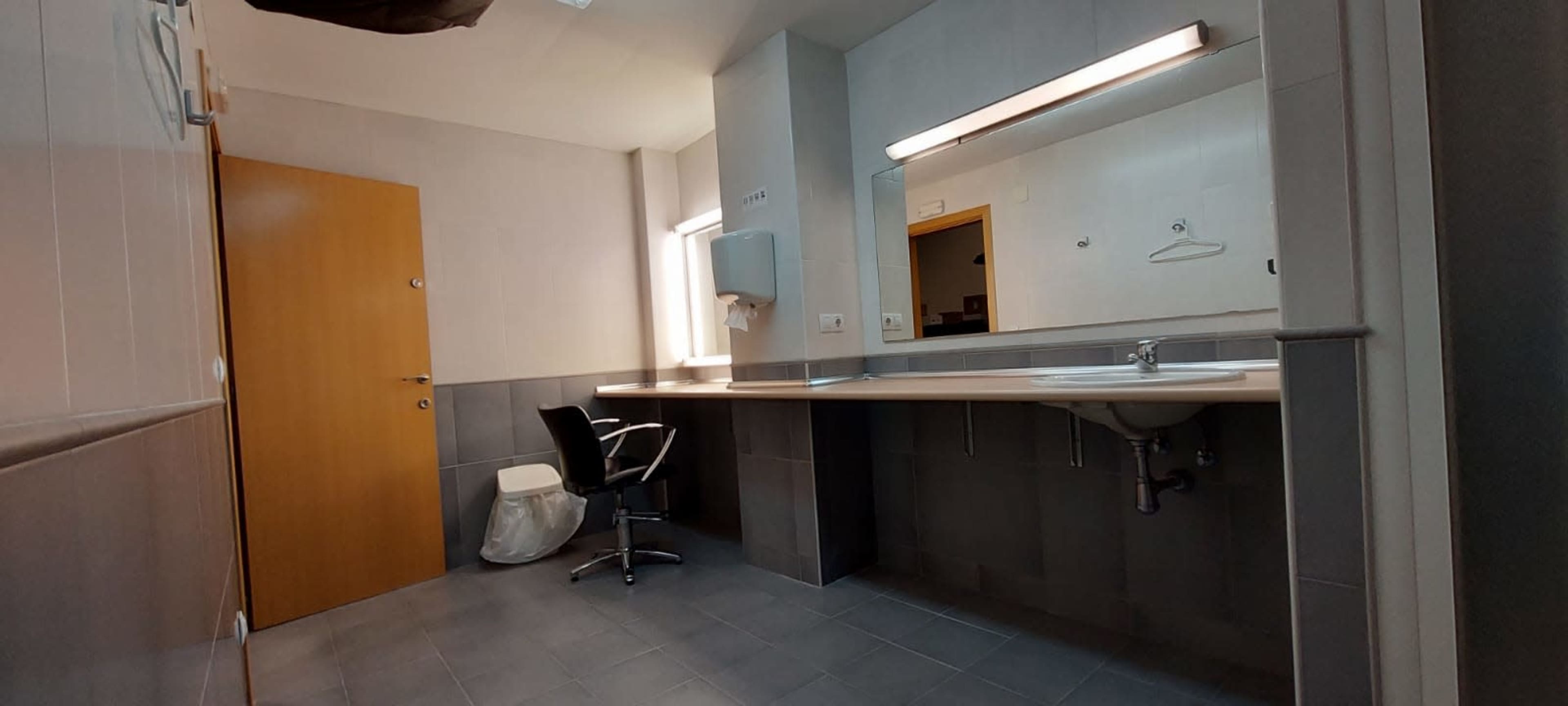 A restroom with a single stall, a mirror above a sink, a chair, and a paper towel dispenser.