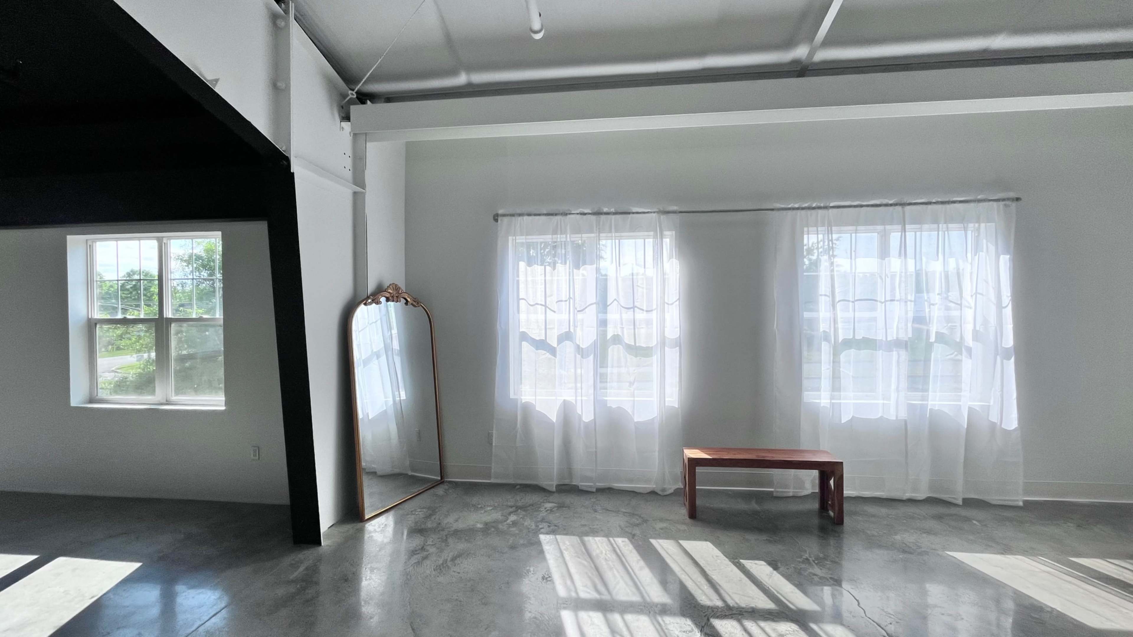 The image shows a room with two large windows covered by sheer curtains, a wooden bench in the center, and a tall mirror leaning against the wall.
