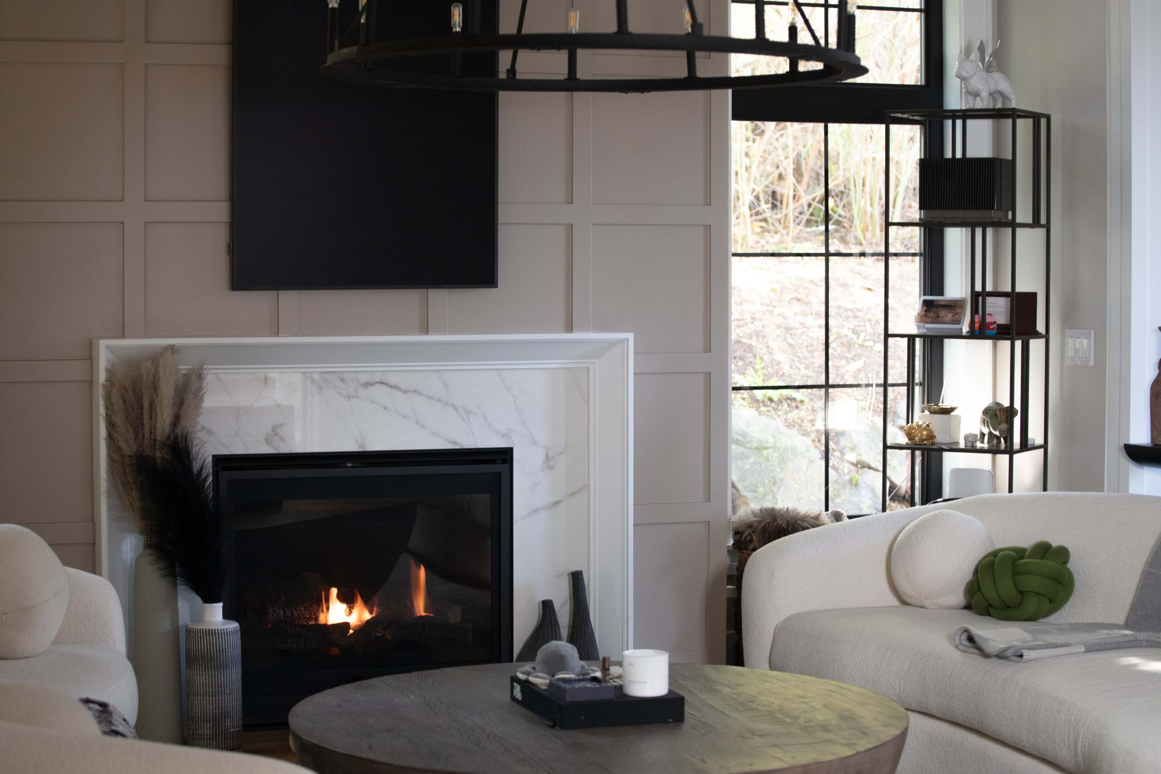 The image shows a modern living room with a fireplace, a round wooden coffee table, and a sectional sofa, all framed by large windows.