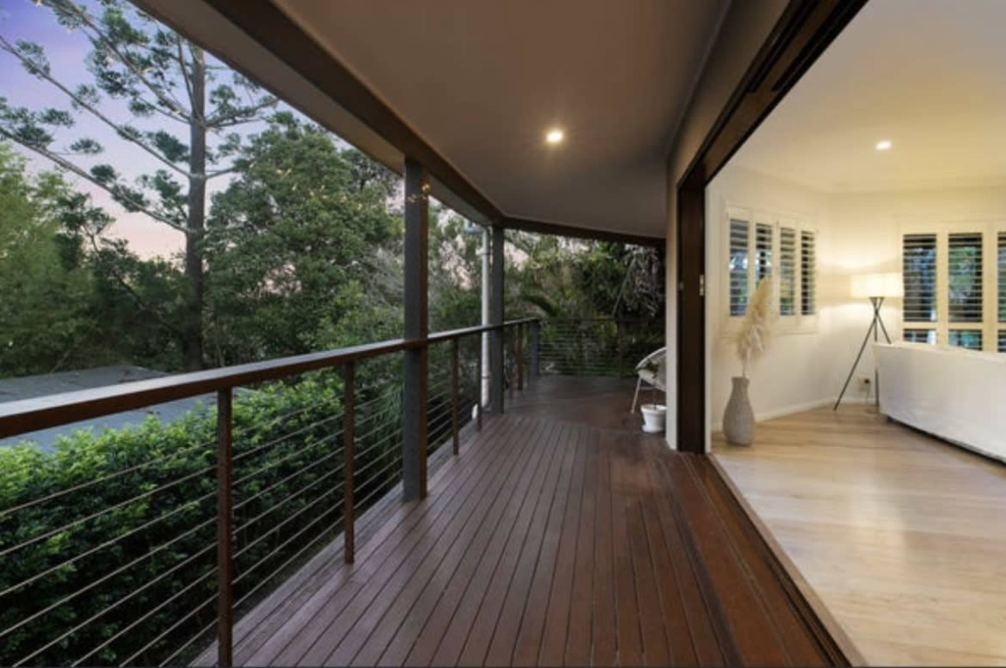 A wooden deck extends from a modern interior space, surrounded by greenery and lit by soft artificial light.