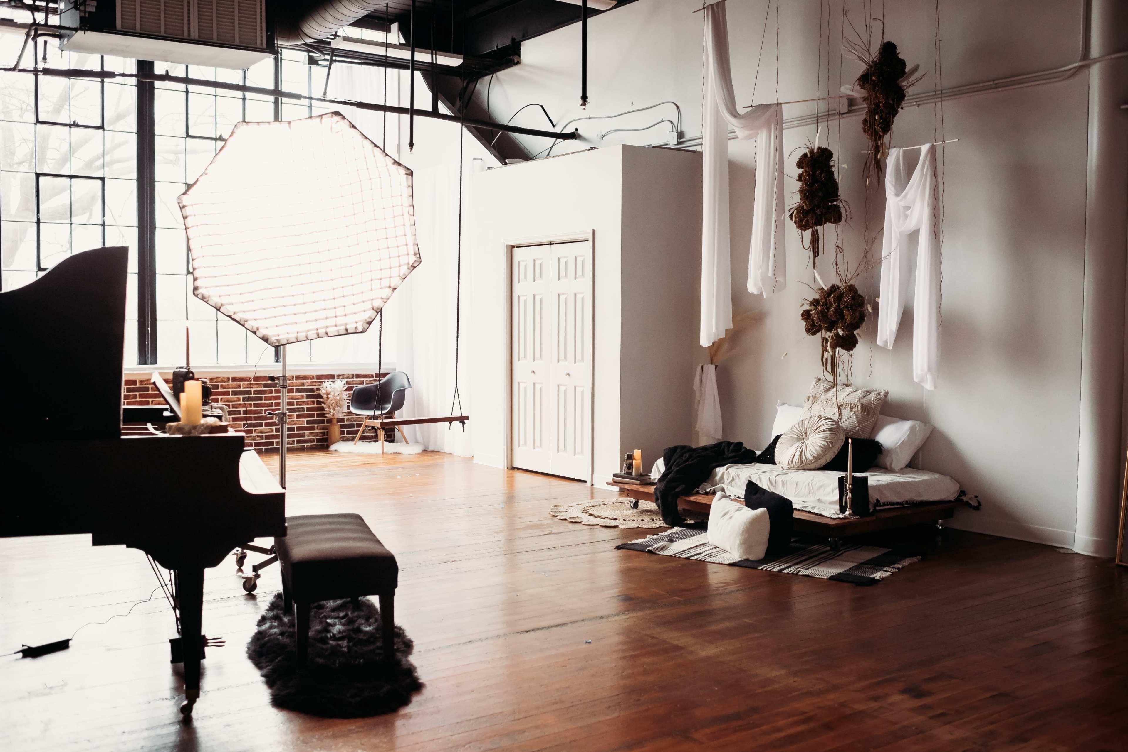 A spacious studio features a grand piano, a bed adorned with various textiles, and soft lighting from a large diffuser near a large window.