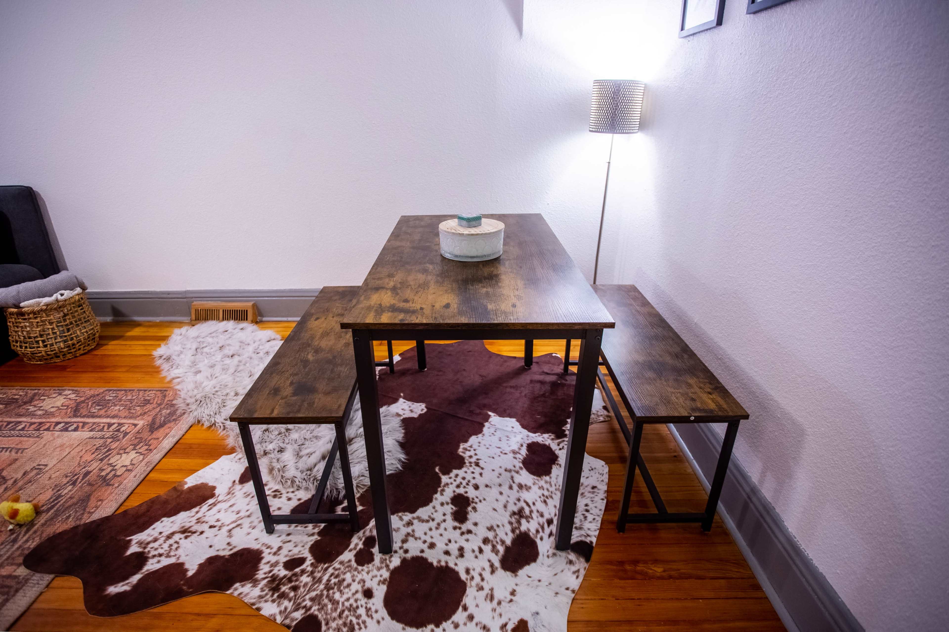 A wooden dining table with two benches is positioned on a cowhide rug in a small room lit by a modern lamp.