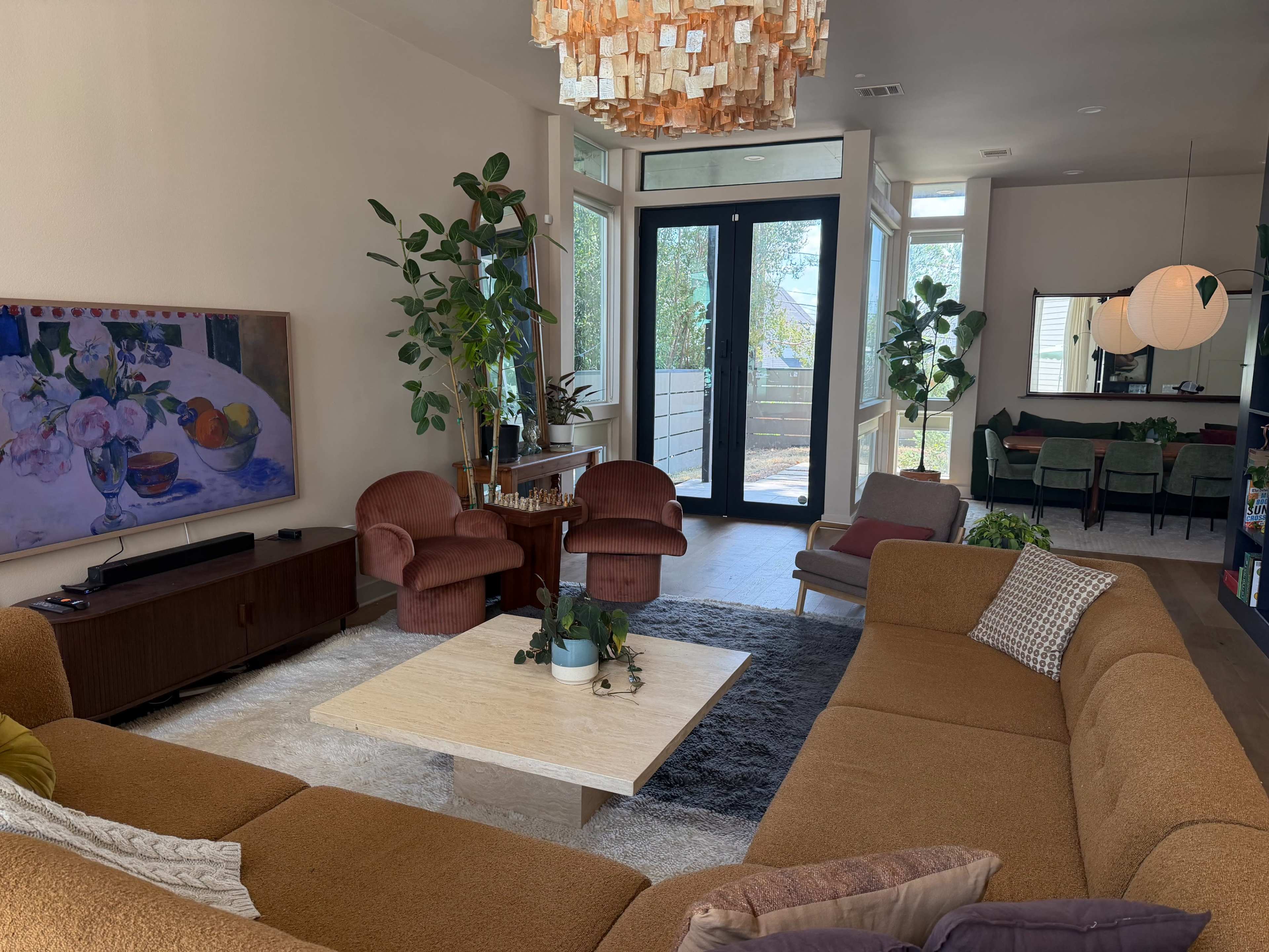 The scene shows a modern living room with a large sectional sofa, low coffee table, potted plants, and glass doors leading to an outdoor area.