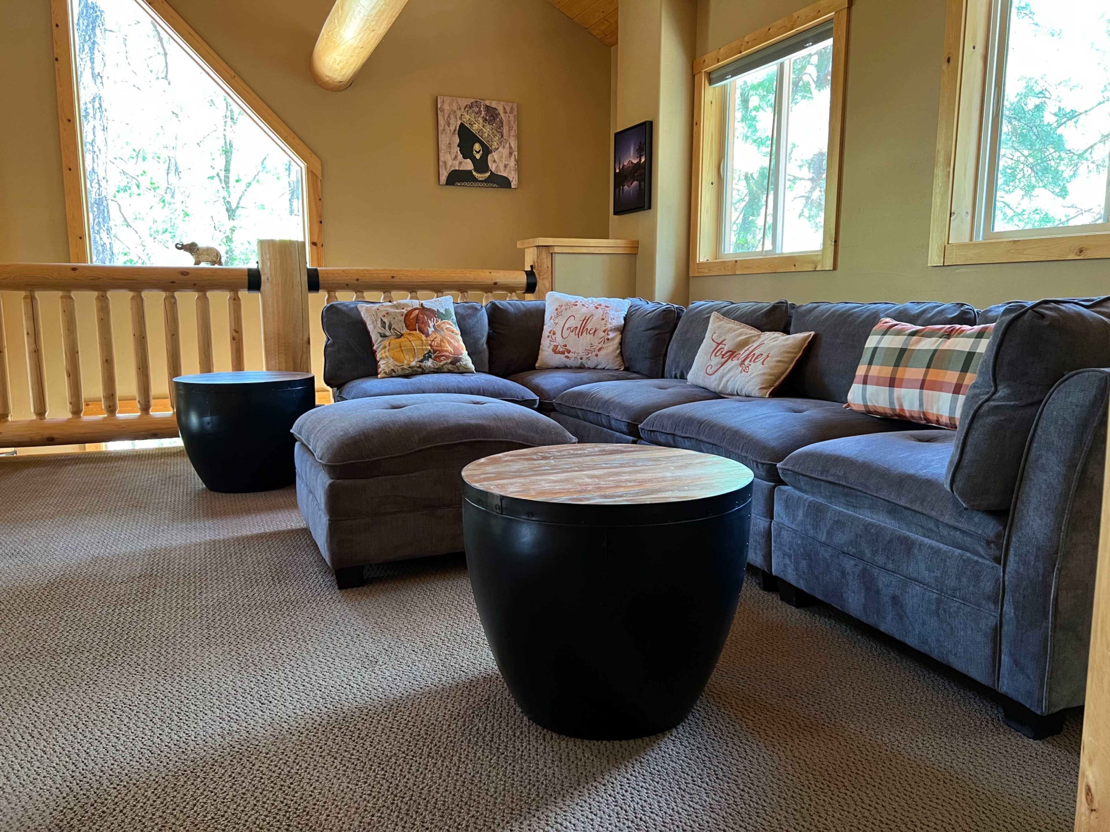 The image shows a cozy living room with a gray sectional sofa, round black coffee tables, and wooden accents, surrounded by large windows that let in natural light.