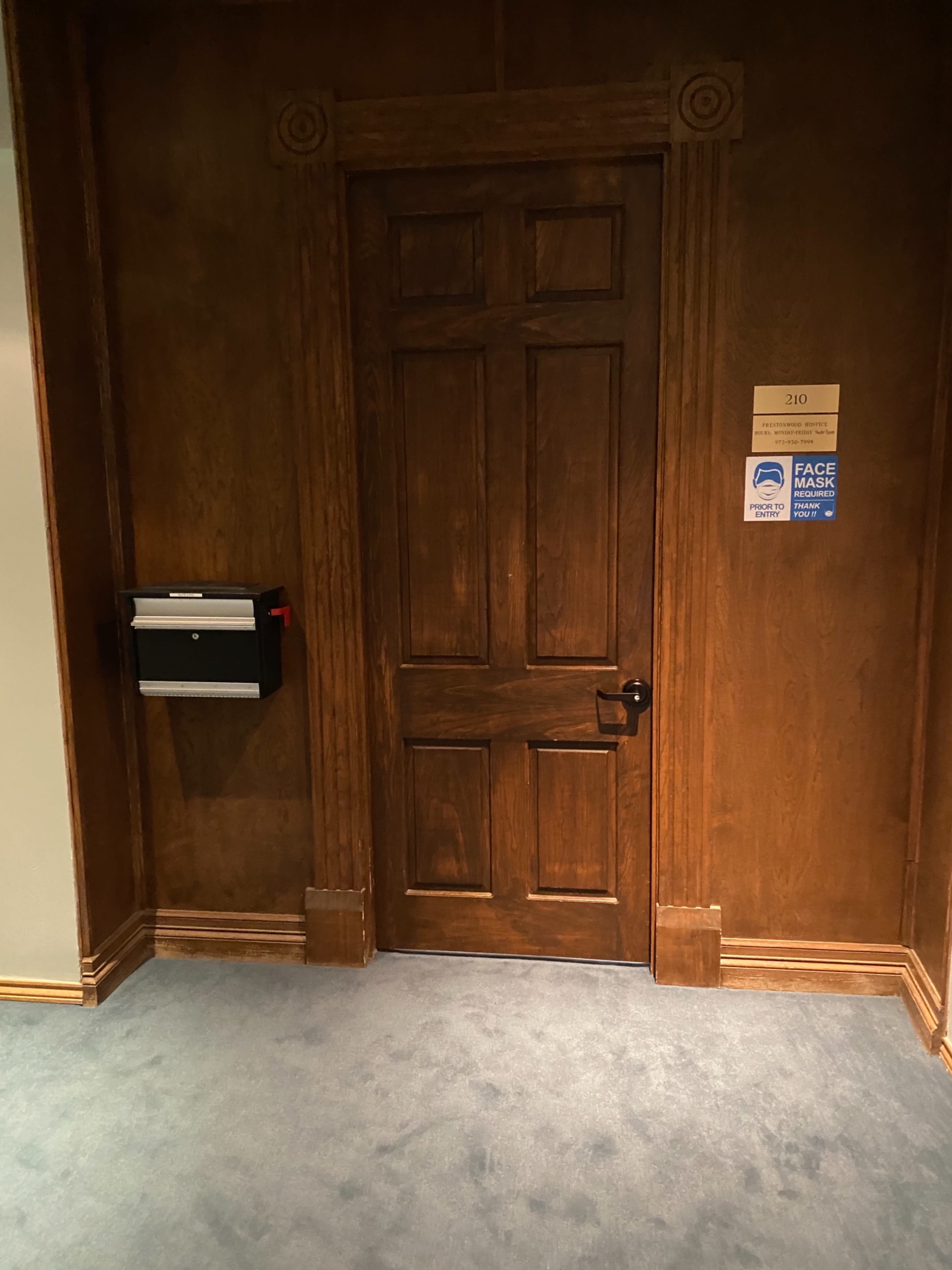 A wooden door marked "210" next to a mailbox on a wall with dark wood paneling and a carpeted floor.
