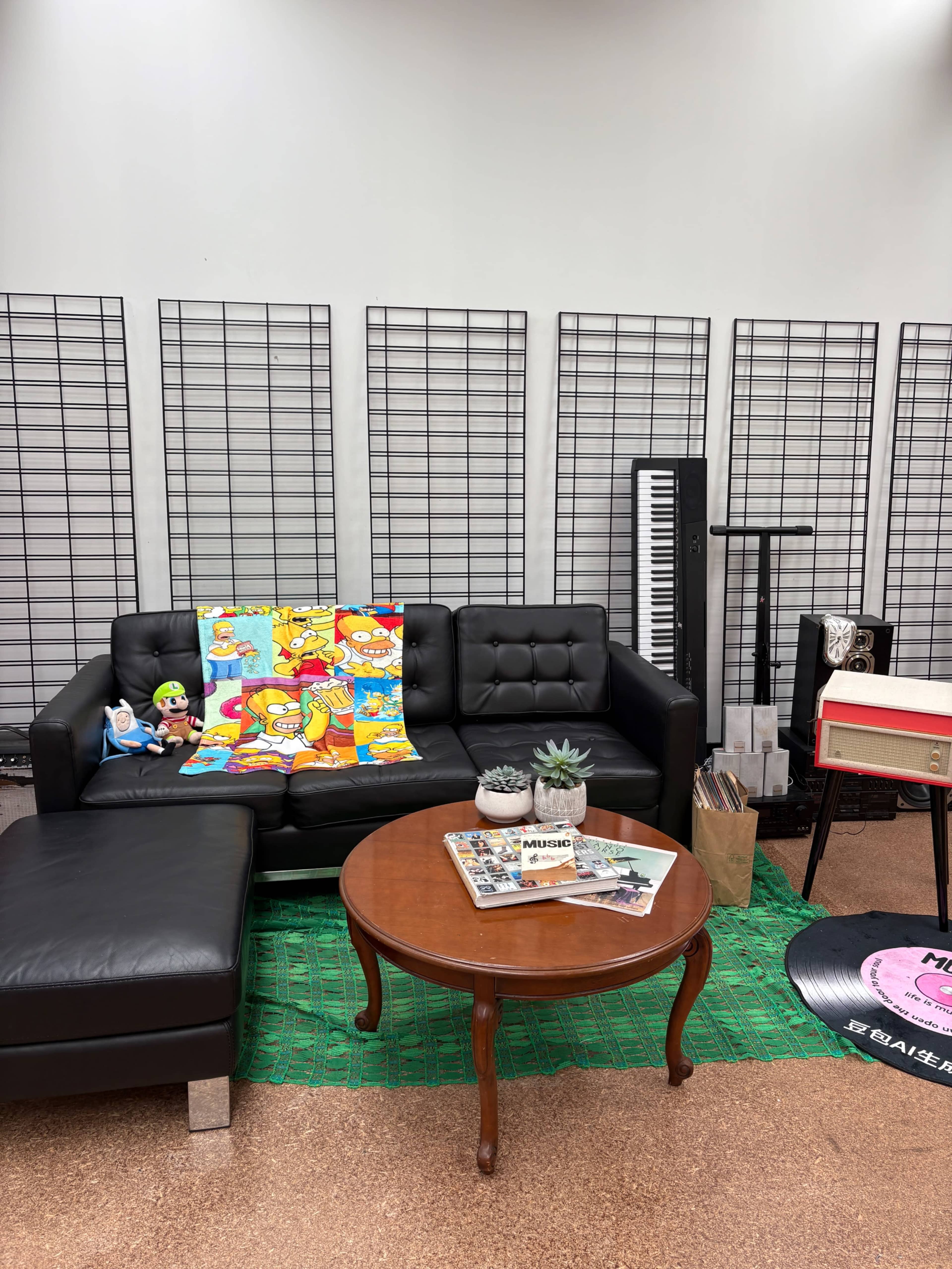 The image shows a cozy lounge area with a black leather sofa, a round wooden coffee table, and various decor items, including colorful blankets, a record player, and a keyboard against a plain wall.