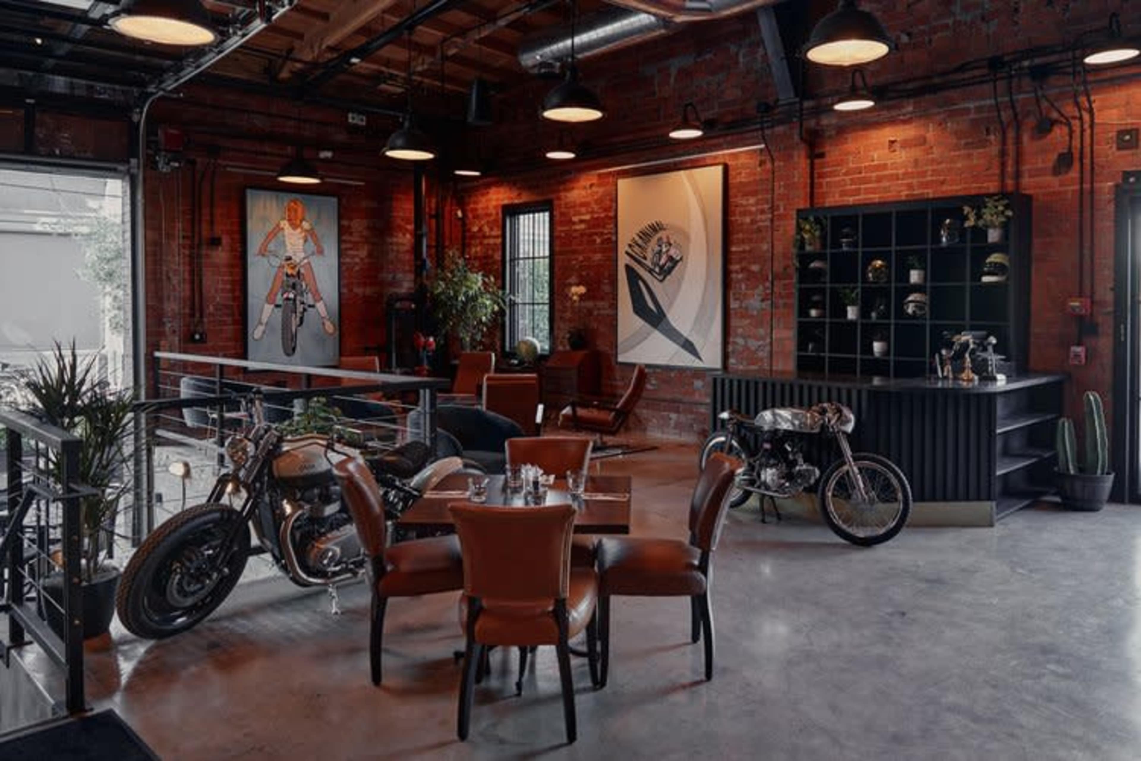 A modern café features industrial decor with exposed brick walls, motorcycle art, and a mix of seating arrangements including chairs and tables.