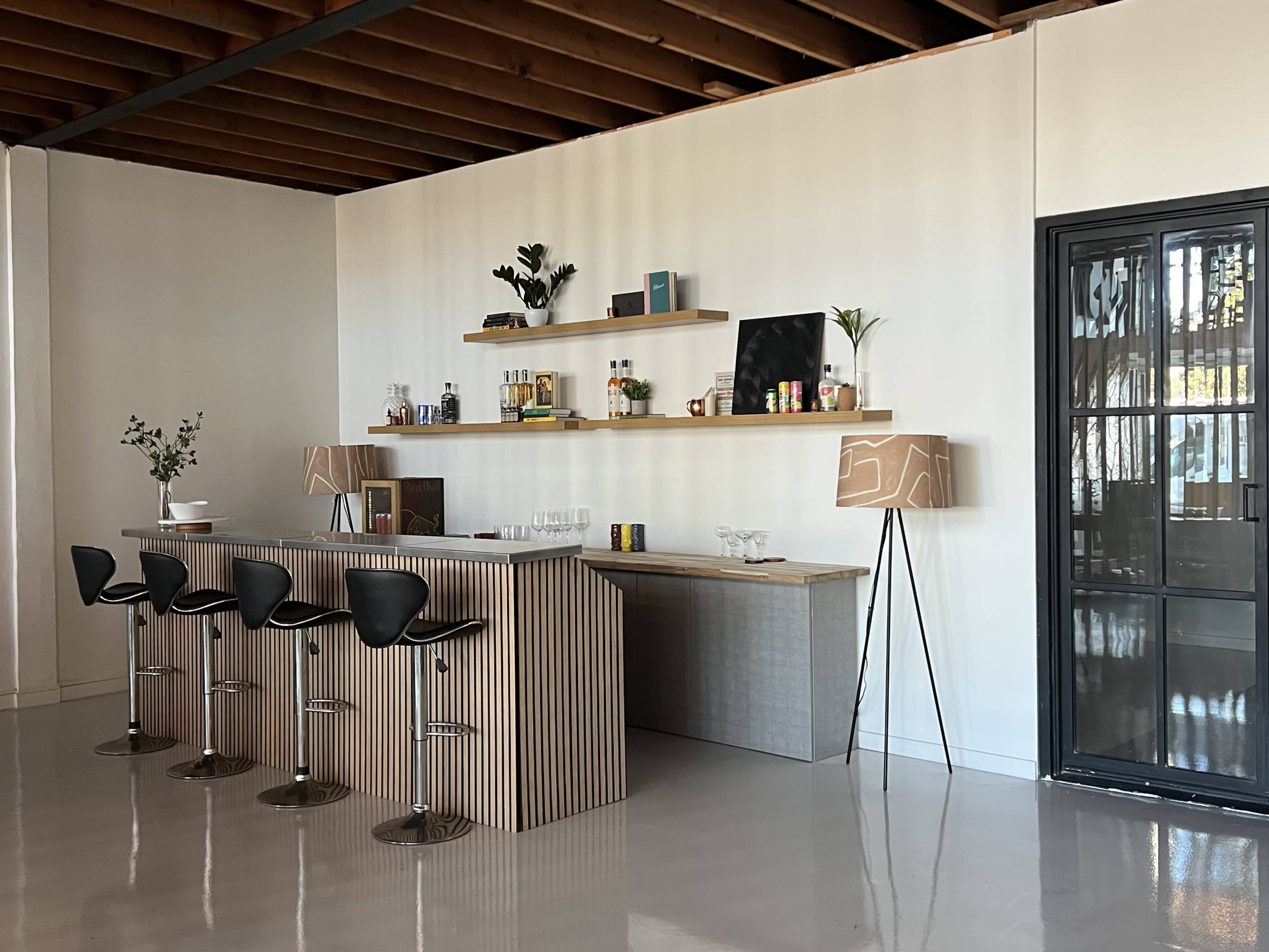The image shows a modern bar area featuring a wooden counter with high stools, a shelf displaying various bottles, and a large glass door.