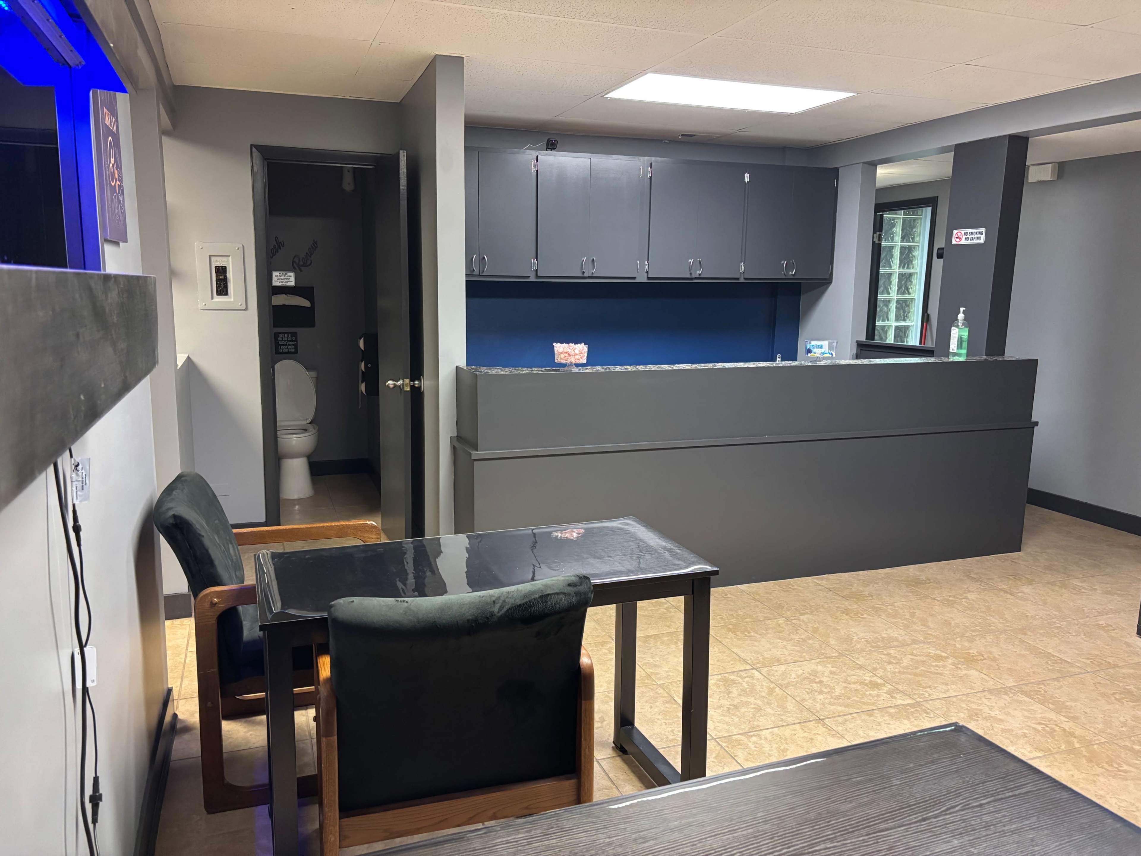 The image shows a modern reception area with a dark gray counter, a table with chairs, and a restroom door visible in the background.