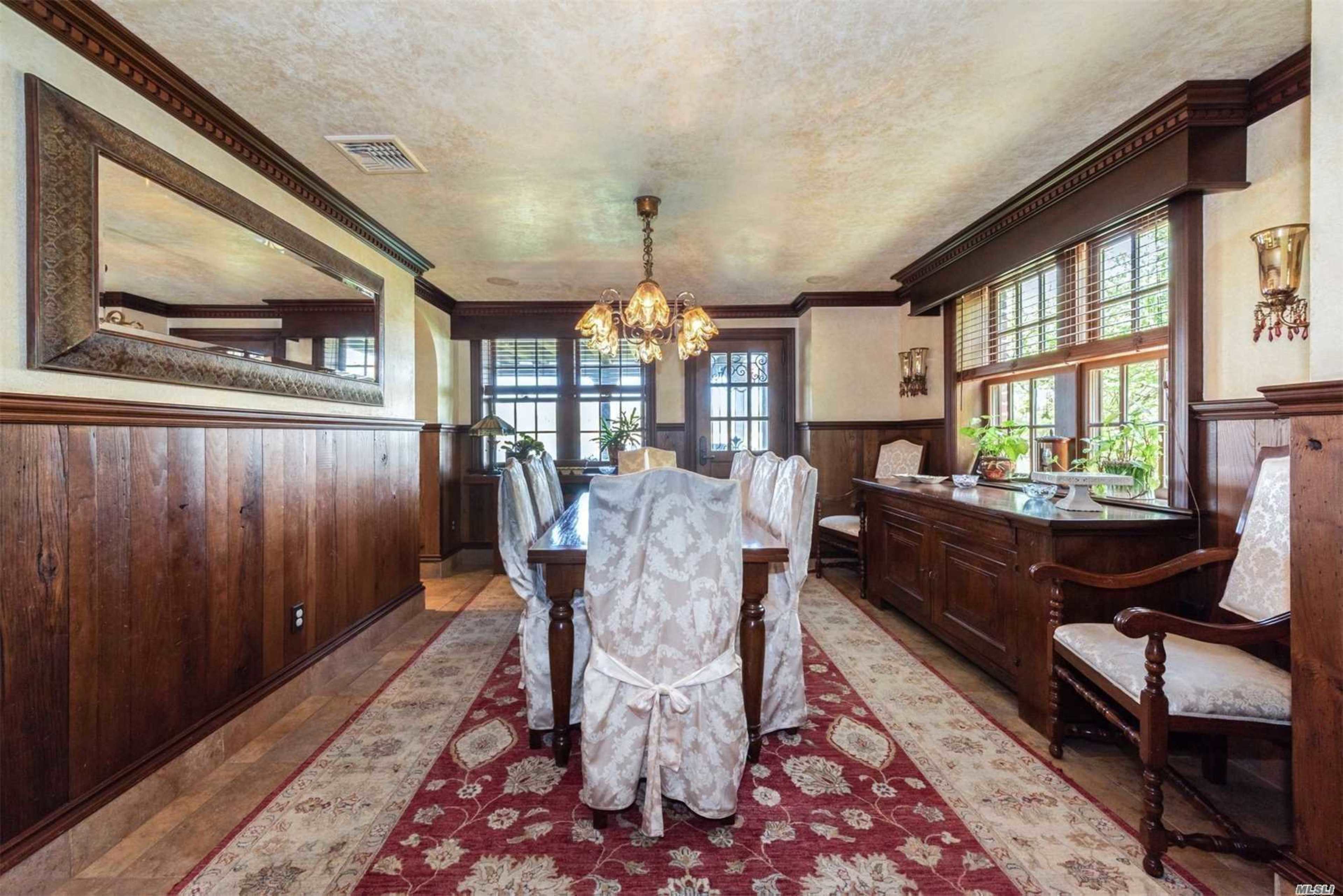 A dining room features a large table surrounded by upholstered chairs, with wood-paneled walls and a chandelier hanging from the ceiling.