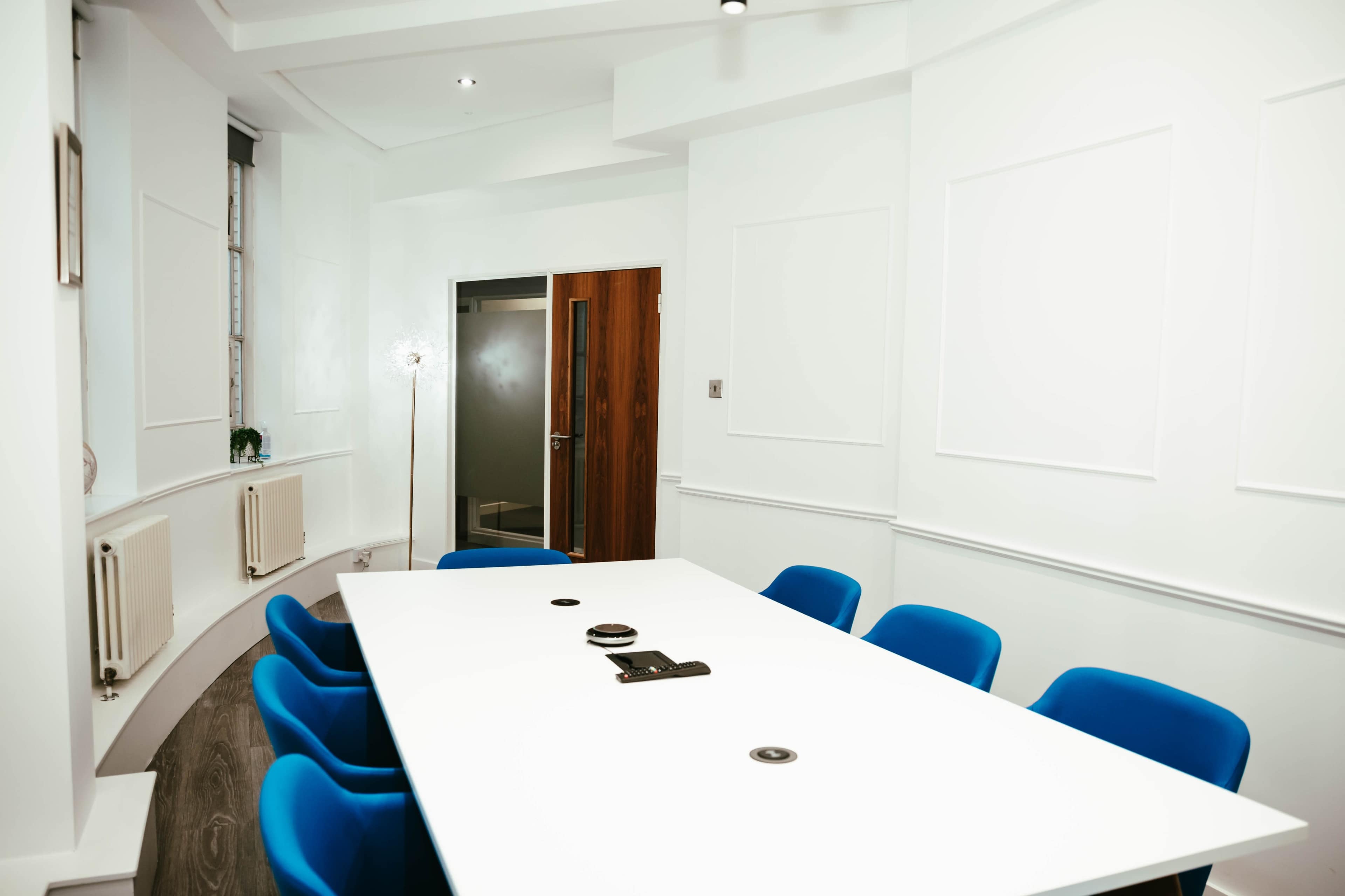 A conference room features a white table surrounded by blue chairs, with a doorway leading to a dark area.