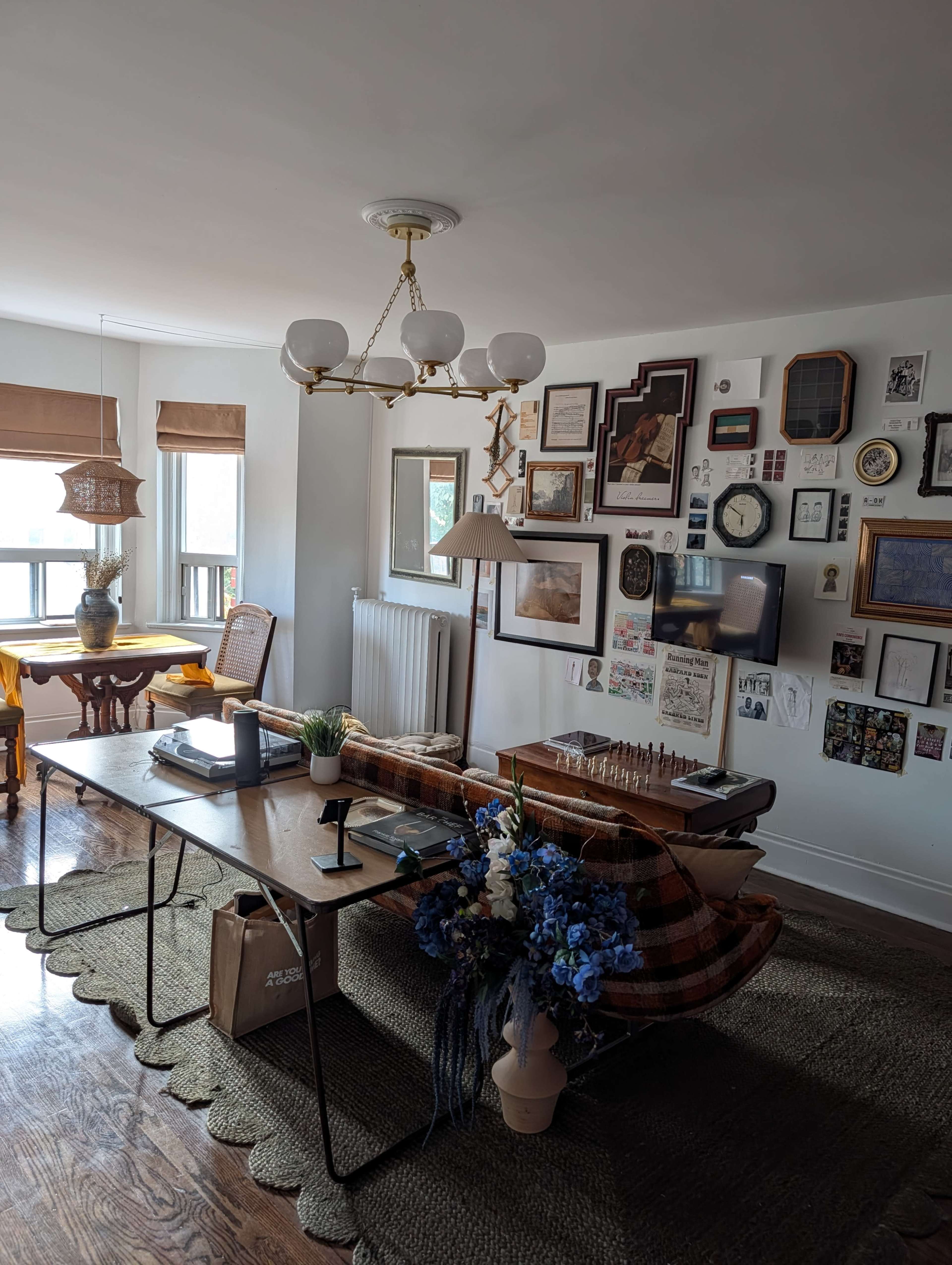 The image shows a bright room with a dining table, a patterned sofa, various wall decorations, and a floral arrangement on a side table.