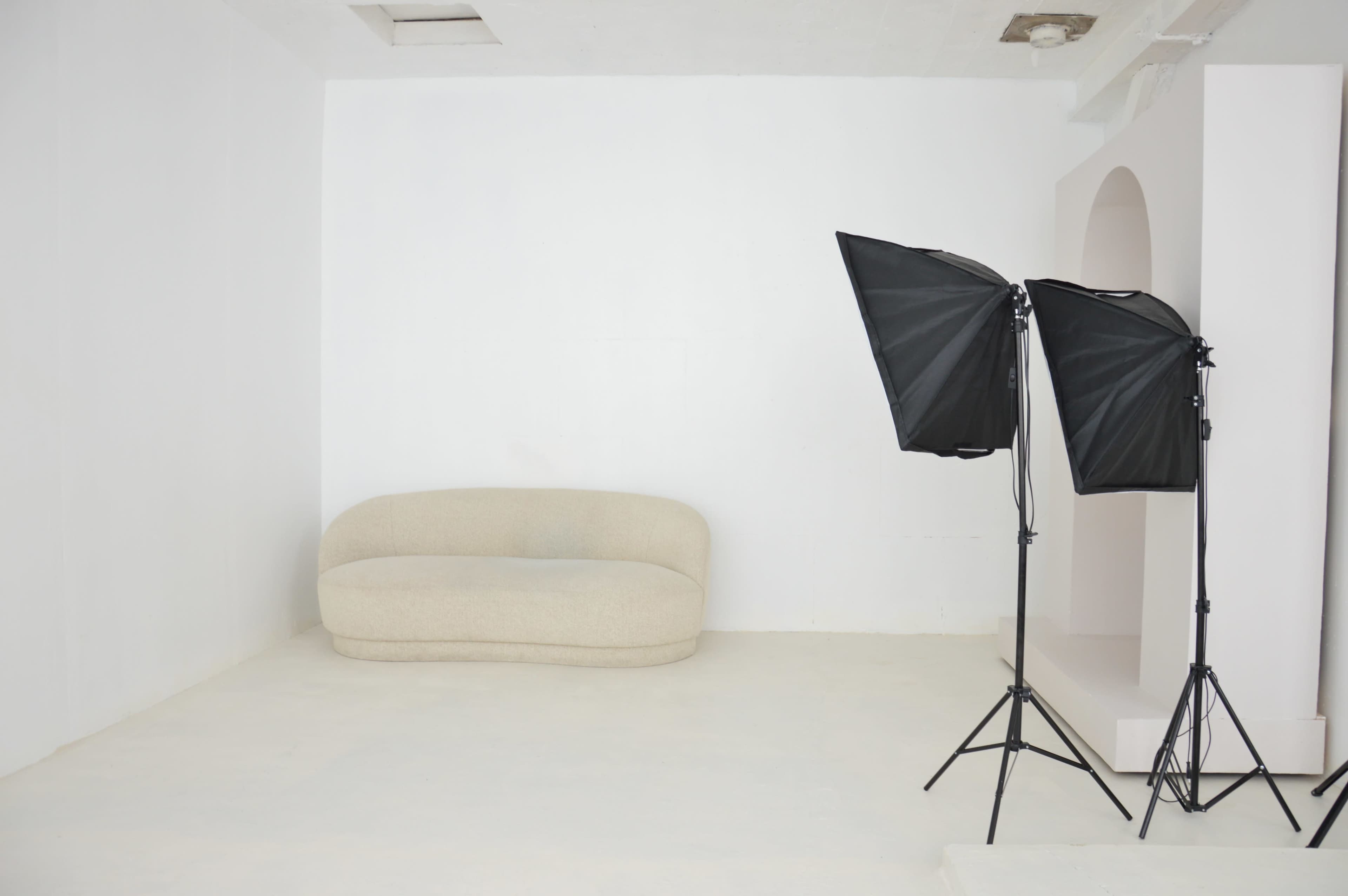 The image shows a minimalist room with a curved beige couch and two black softbox lights on stands.