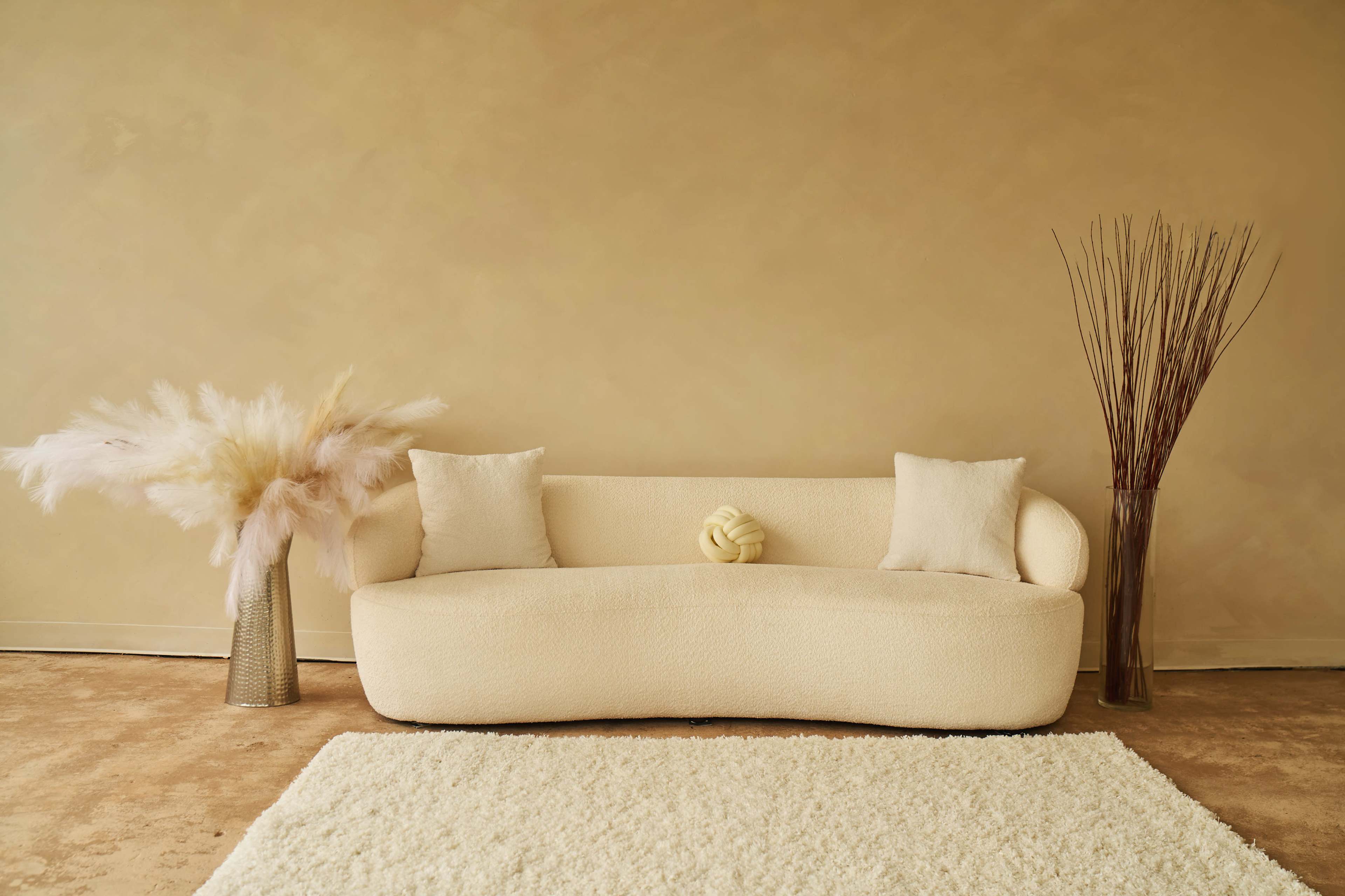 A light-colored, sculptural sofa with two pillows sits against a textured beige wall, accompanied by a vase with dried grasses and a fluffy rug on the floor.