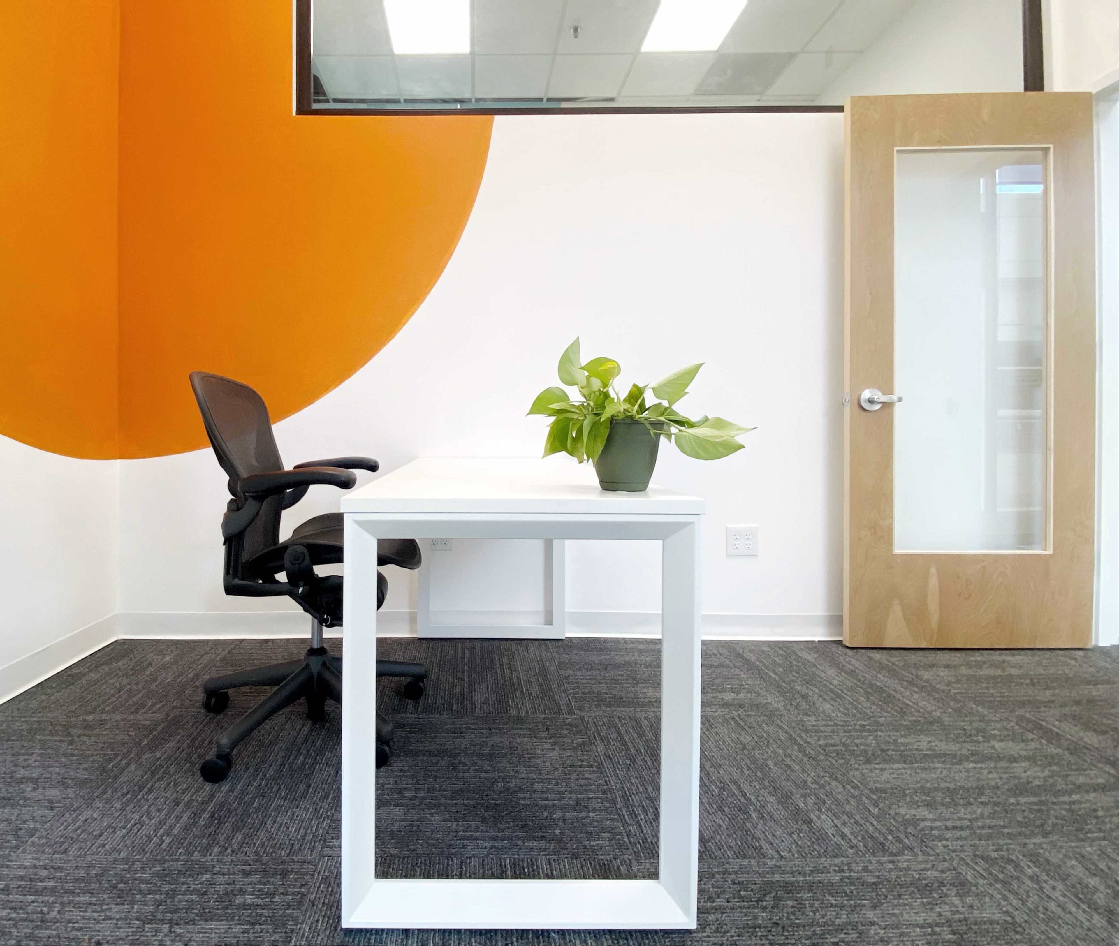 A minimalist office features a white desk and a black chair beside a potted plant, with an orange accent wall and a door in the background.