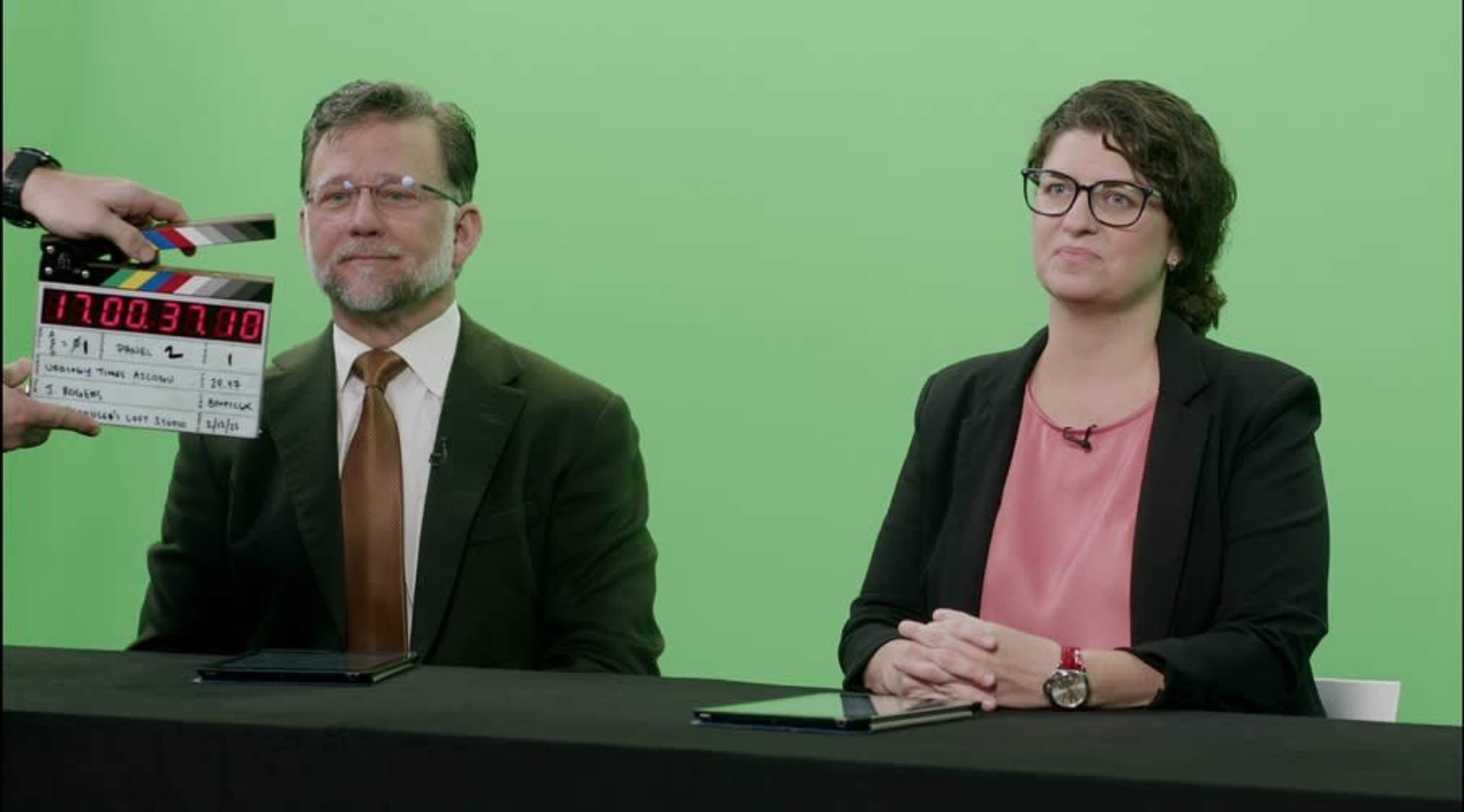 Two individuals sit at a black table in front of a green screen, with one person holding a clapperboard.