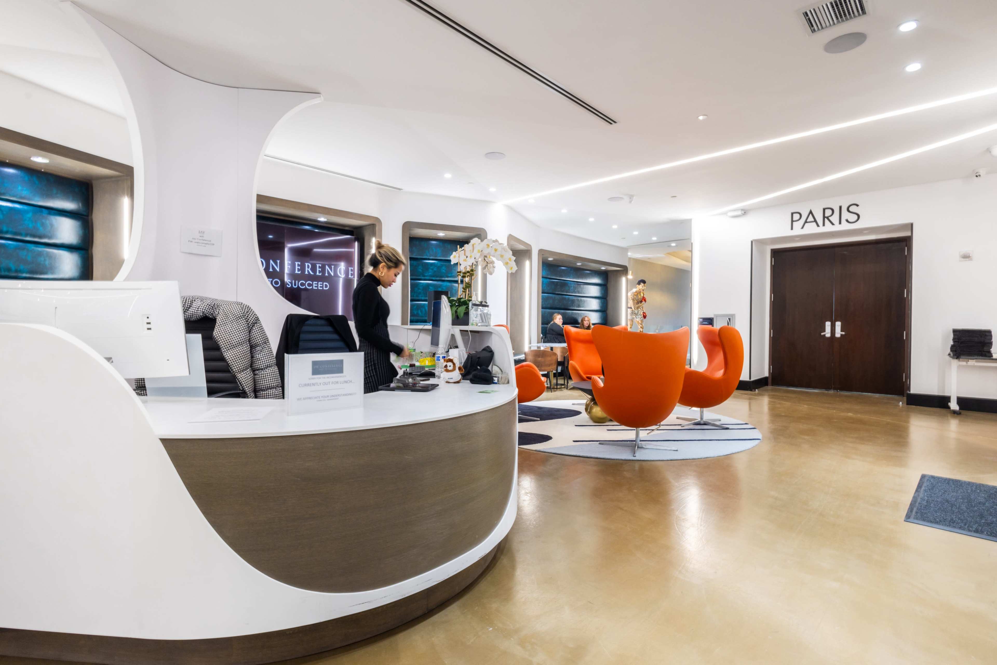 A modern reception area with a white curved front desk, orange seating, and a sign indicating "PARIS" on a wall.
