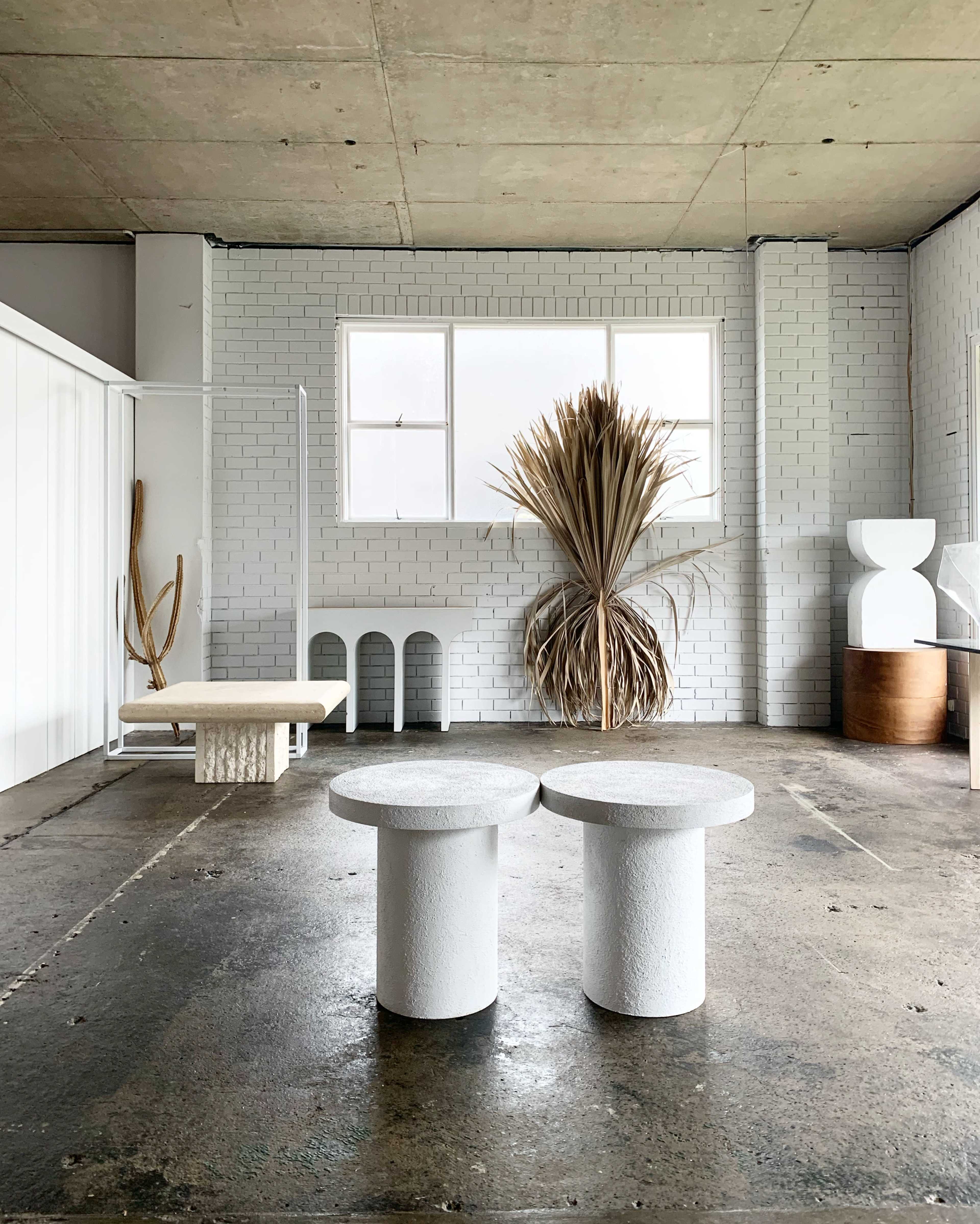 A minimalist interior space with two round white tables in the foreground and a large dried plant arrangement near a window.
