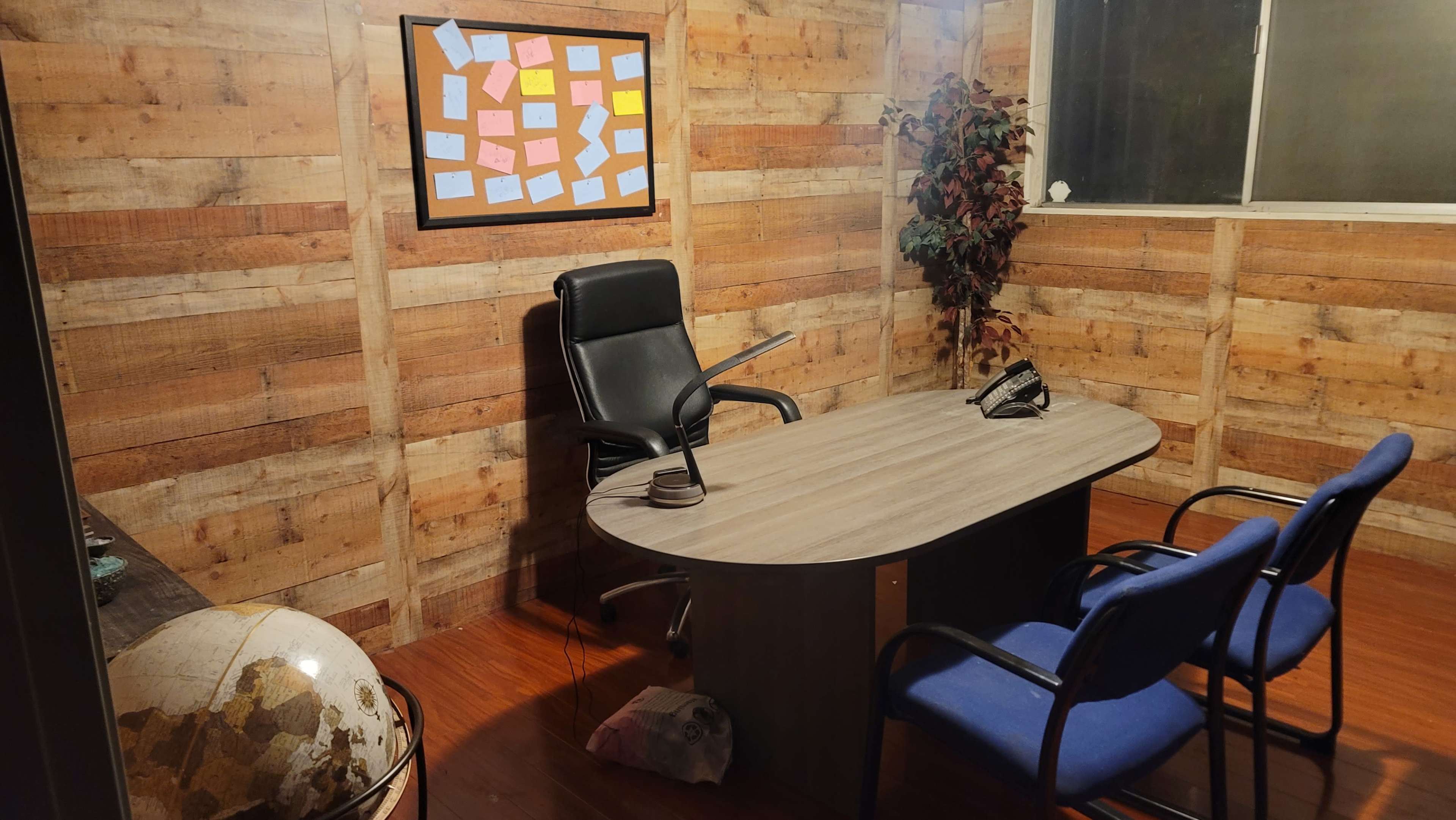 A small office with a round desk, a black chair, two blue chairs, a telephone, a framed board with sticky notes on the wall, and a globe in the corner.