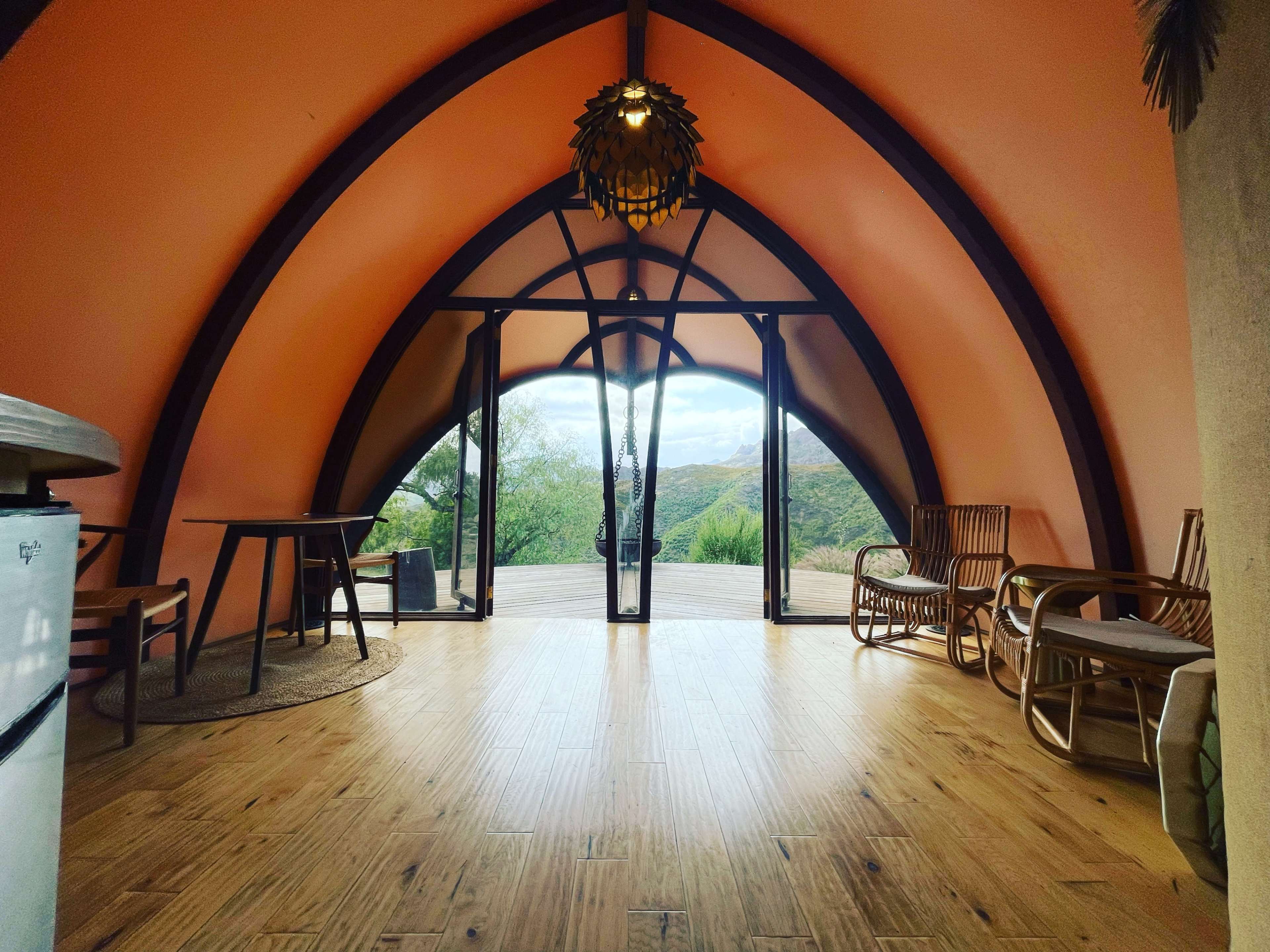 The interior of a curved, modern structure features orange walls, wooden flooring, and large glass doors that offer a view of the surrounding landscape.